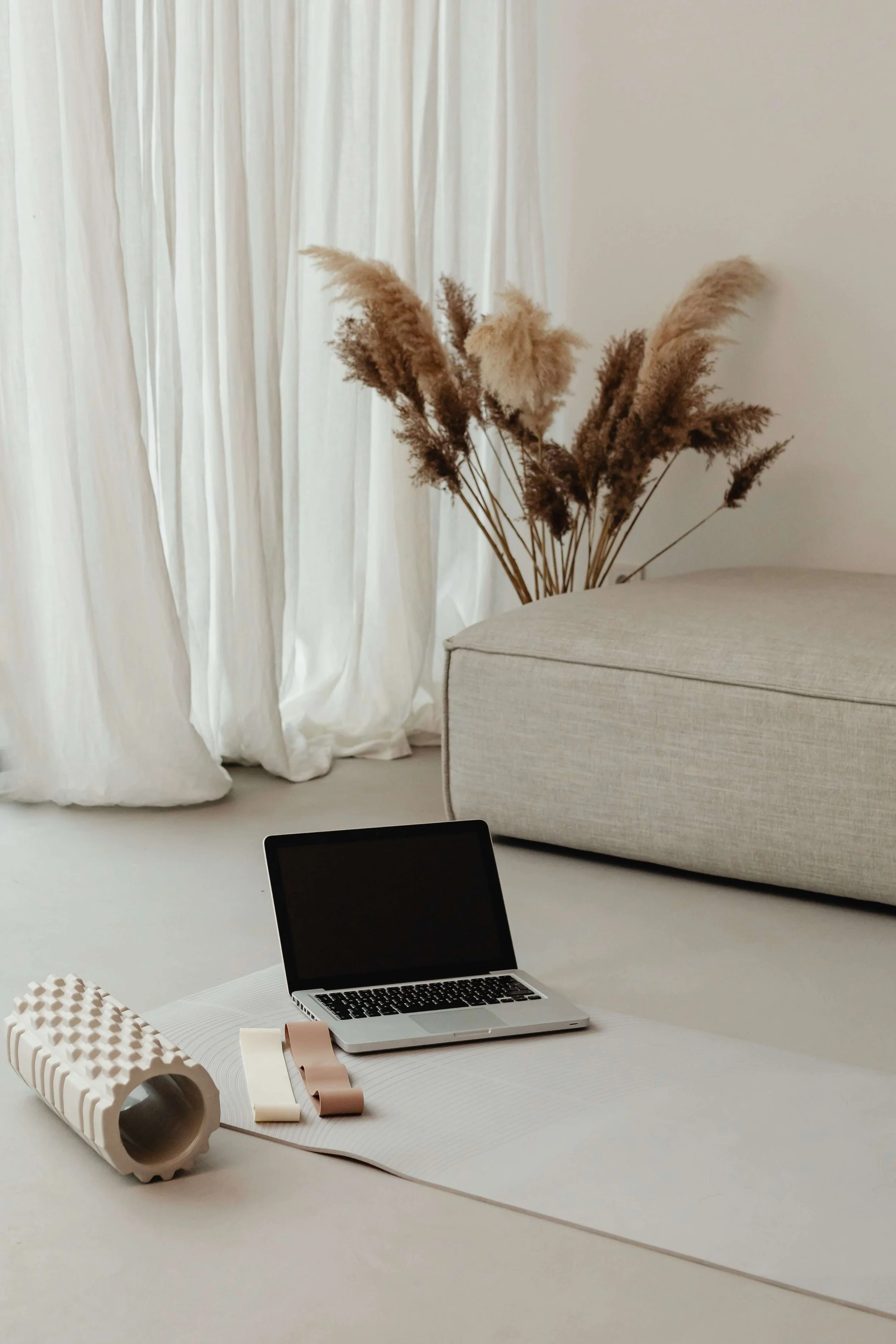 A laptop, foam roller, and resistance bands on a yoga mat in a minimal living room with beige couch, pampas grass, and white curtains.