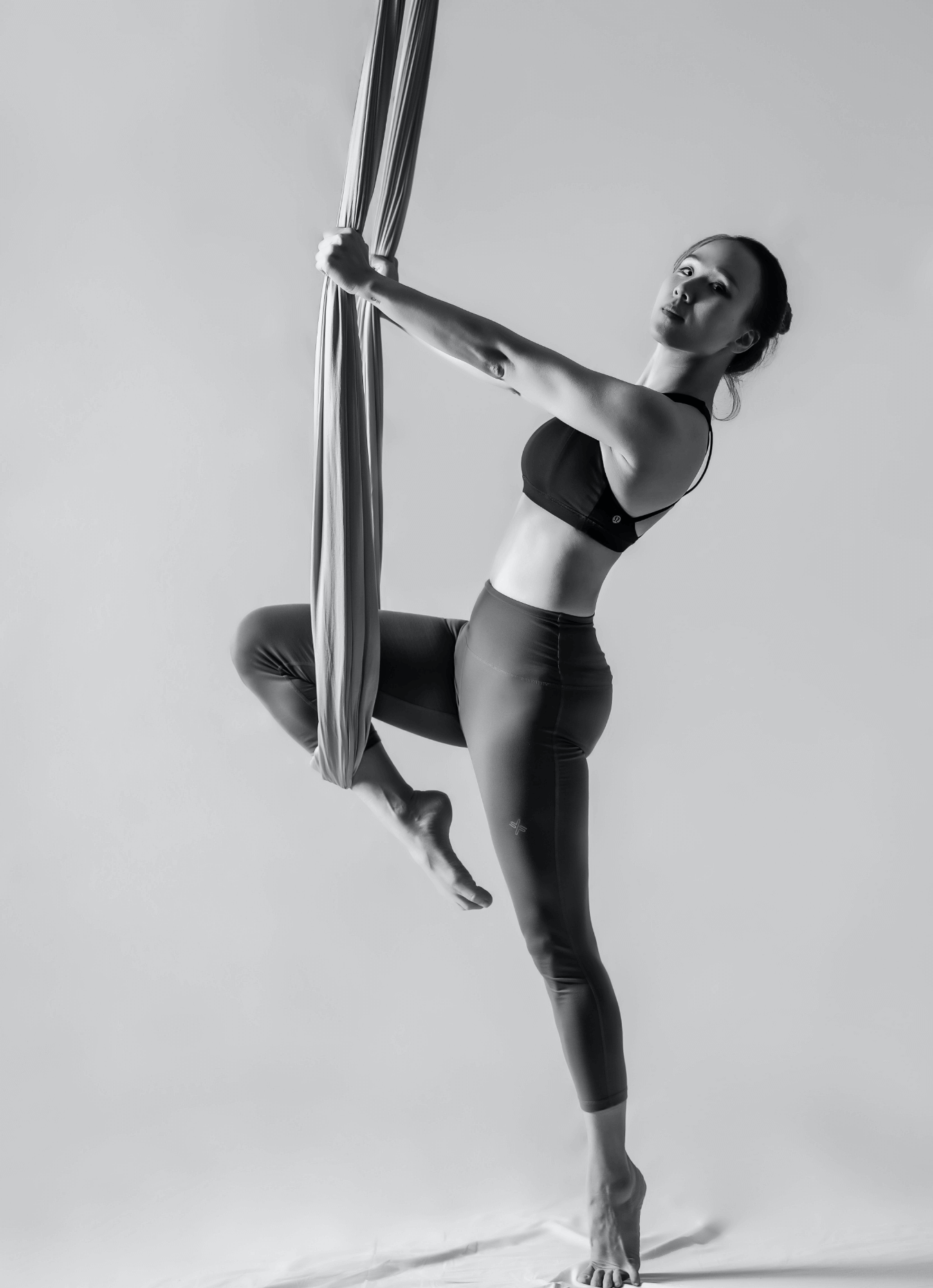 A woman performing aerial silks dance, suspended in the air with one leg bent, holding onto fabric with one hand, wearing a sports bra and leggings, against a plain background.