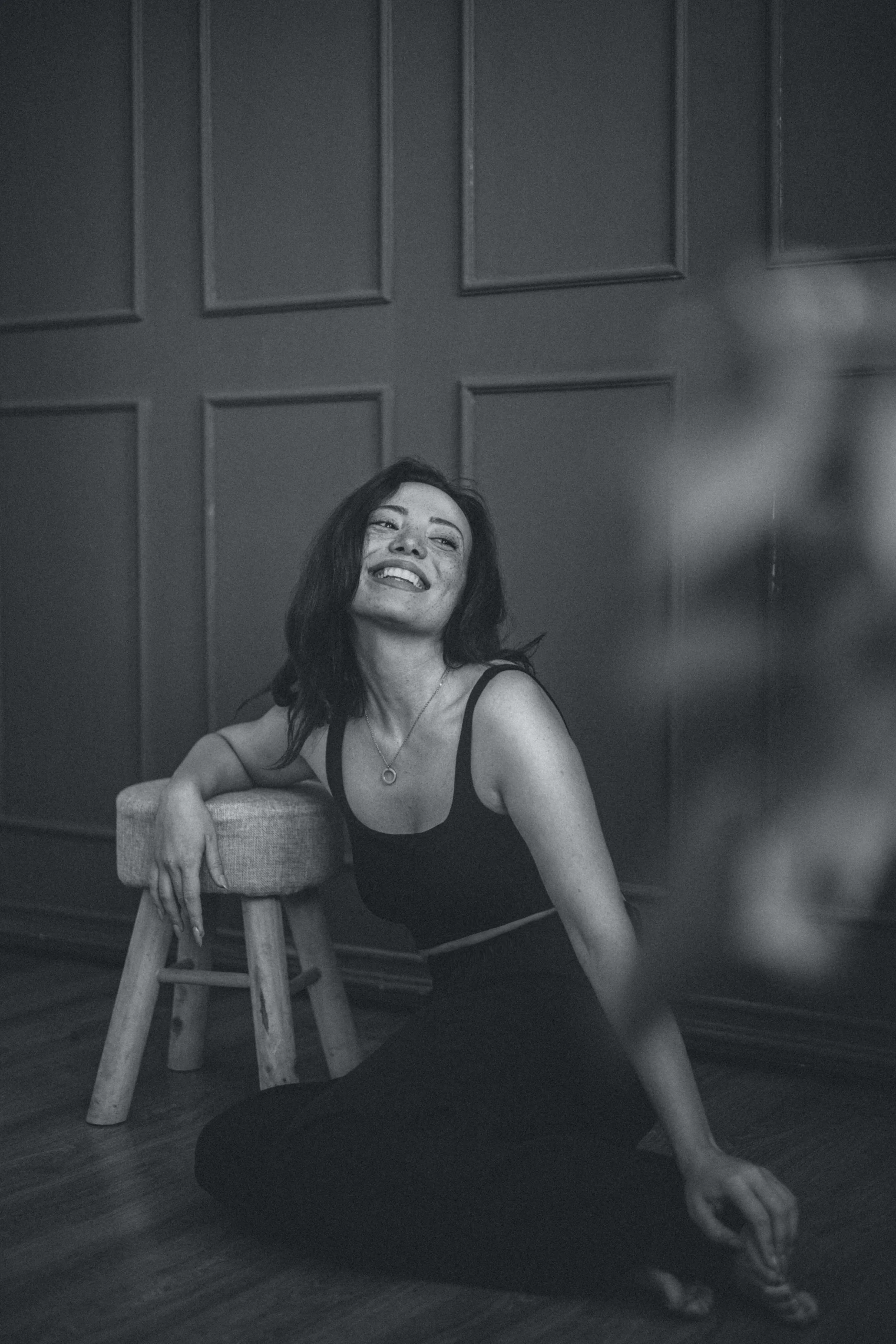 Black-and-white photo of a woman sitting on the floor with her back against a small stool, smiling and looking upward. She has dark hair, freckles, and is wearing a tank top, necklace, and dark pants. The background features a paneled wall.