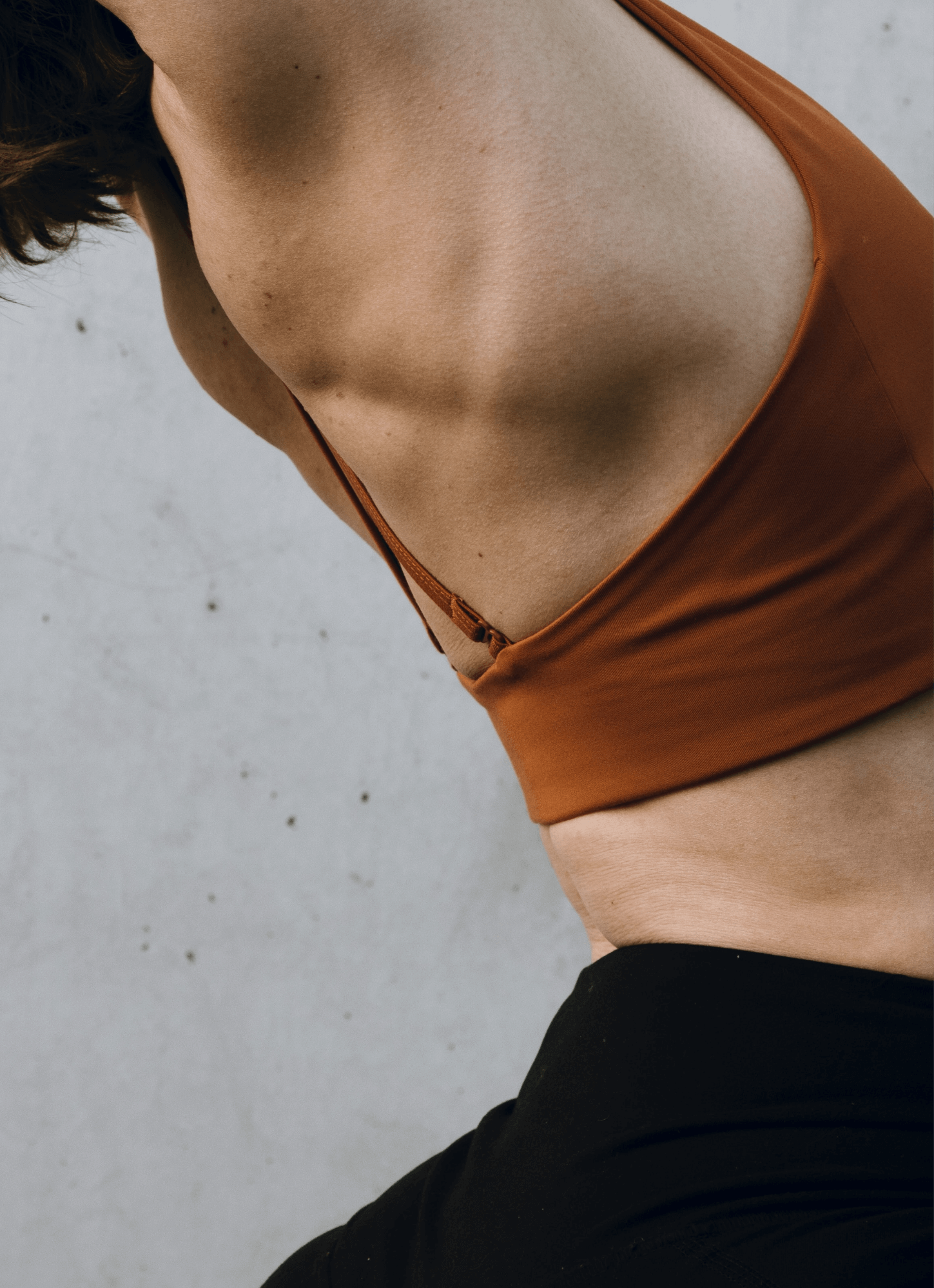 Close-up of a woman's torso wearing a rust-colored sports bra and black bottoms, standing against a plain gray wall.