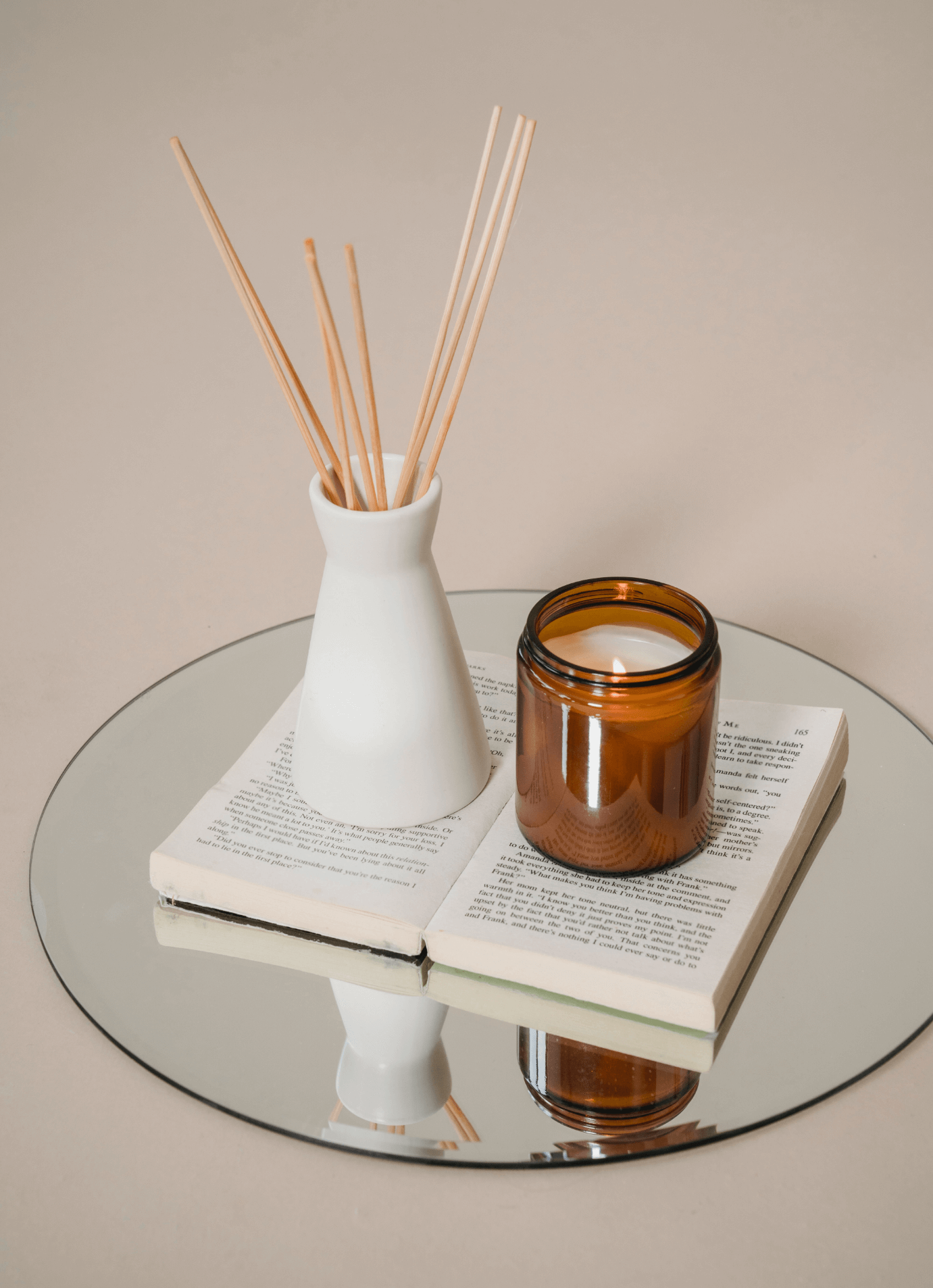 A mirror on a table reflects a white vase with wooden skewers, an open book, and an amber candle jar.