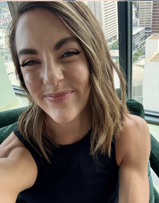 A woman with shoulder-length light brown hair, smiling with slightly squinted eyes, taking a selfie inside a high-rise building. Behind her, large windows reveal a cityscape with tall buildings.