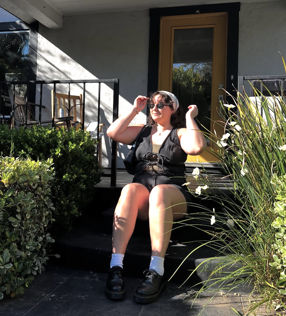 A woman with dark hair, sunglasses, and a black sleeveless top sits on the steps of a porch with greenery around her, adjusting her sunglasses during a sunny day.