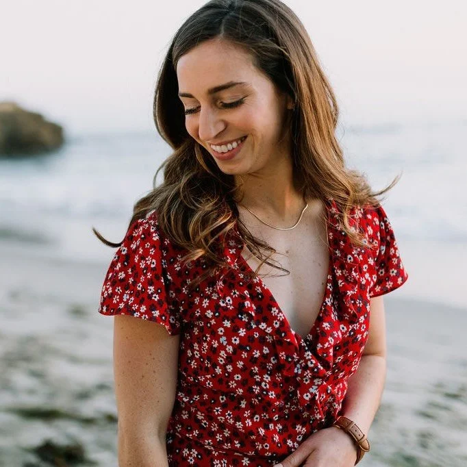 A woman standing on the beach, smiling with her eyes closed, wearing a red floral dress.