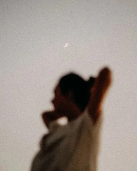 A person standing outdoors with arms raised, looking up at the night sky with a crescent moon visible.