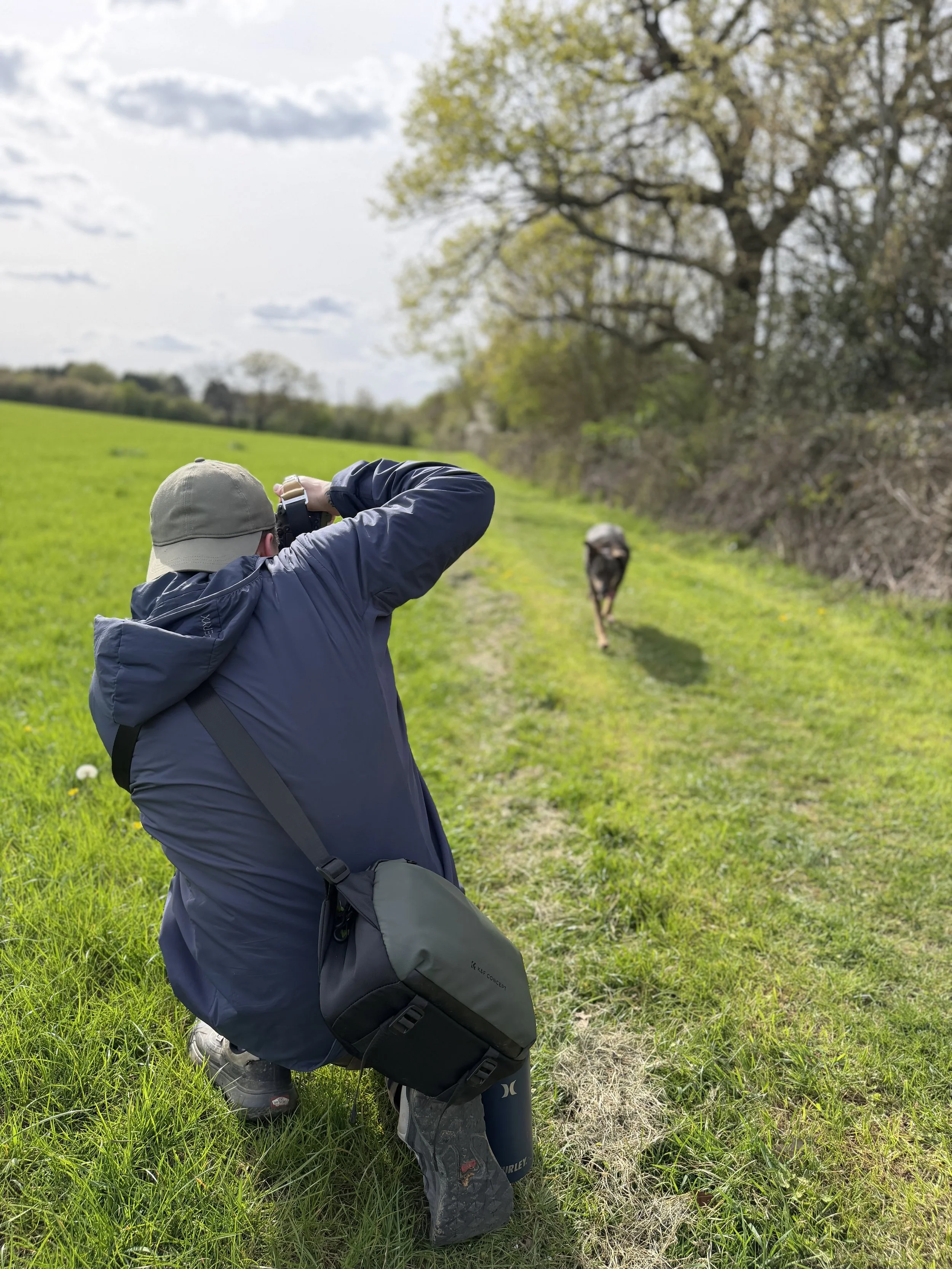 Person kneeling on grass with a camera, wearing a hat, jacket, and carrying a bag, outdoors in a grassy area with trees and a cloudy sky in the background, and a dog approaching in the distance.