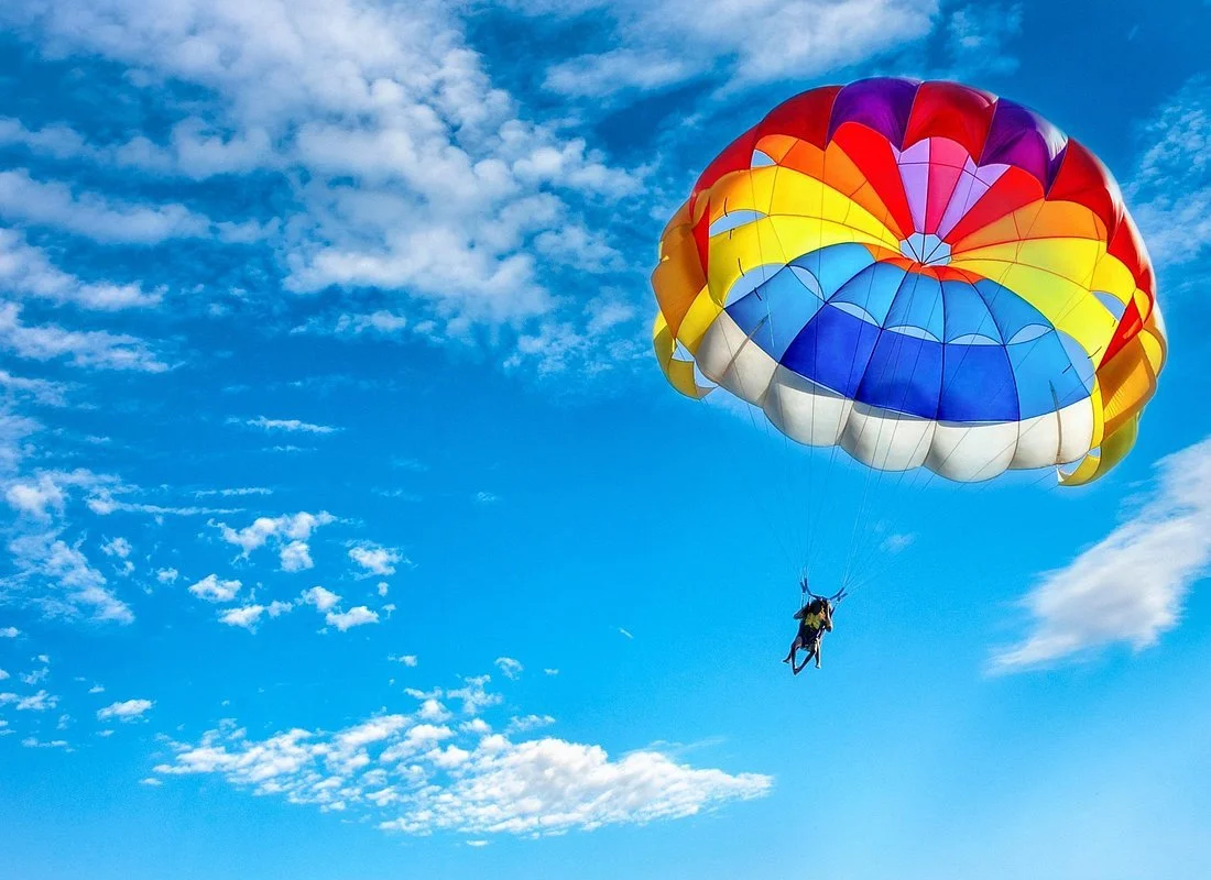 Person parasailing with a colorful parachute in a blue sky with scattered white clouds.