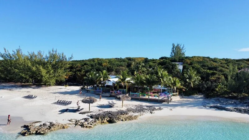 A tropical beach with white sand, clear blue water, palm trees, and a beachside bar or restaurant with umbrellas and lounge chairs.