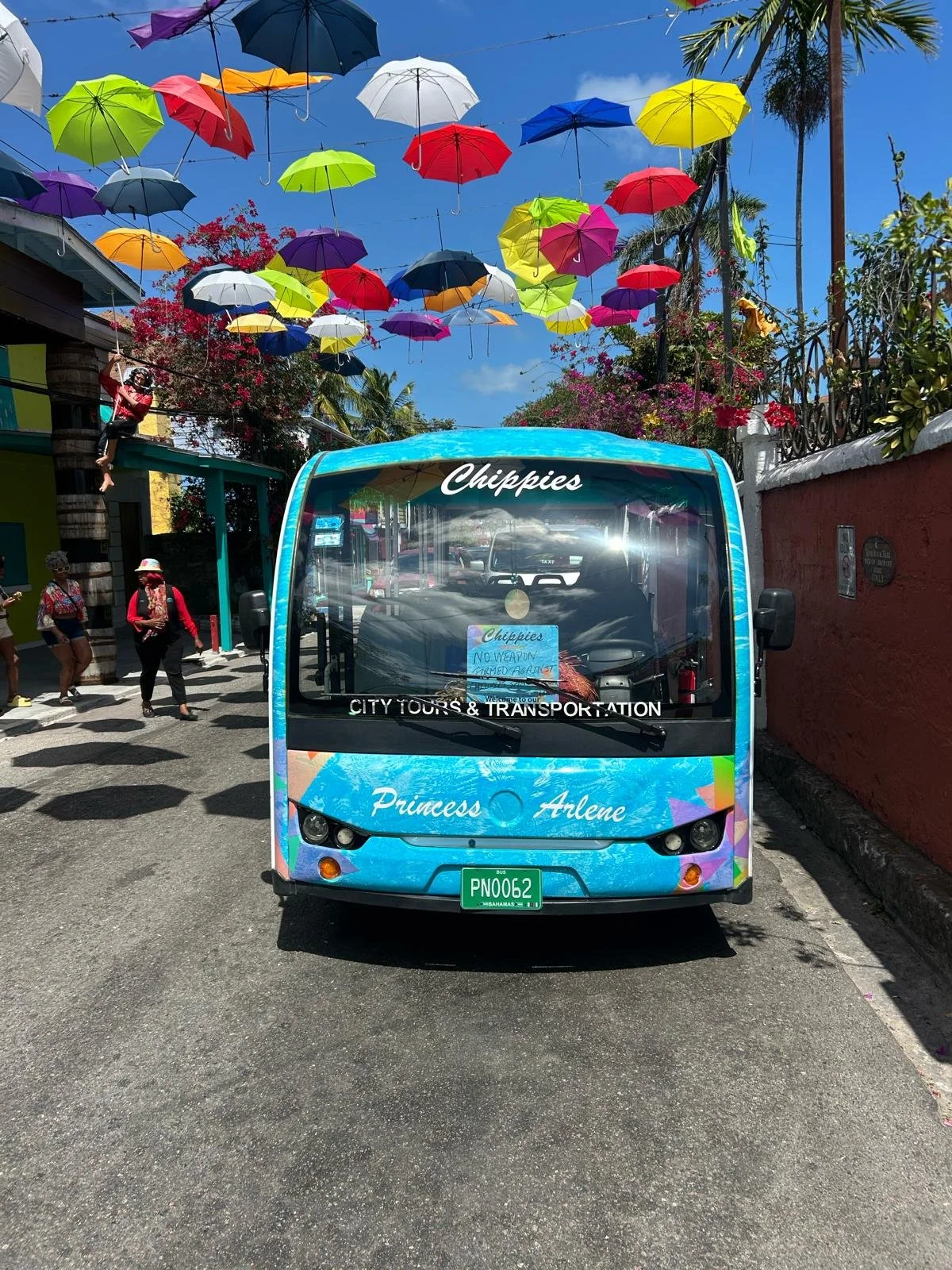 Colorful umbrellas suspended overhead on a street with sightseeing bus in front, pink and red flowers, and tropical trees.
