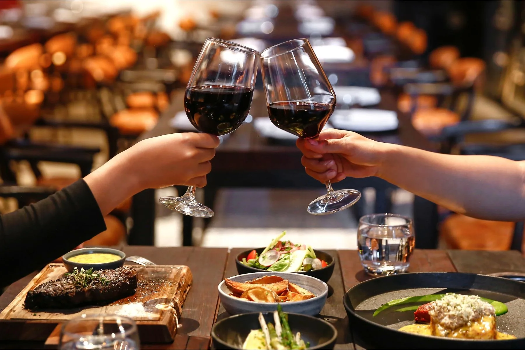 Two people toasting with glasses of red wine at a dinner table filled with various dishes, including steak, salad, and vegetables in a cozy restaurant setting.