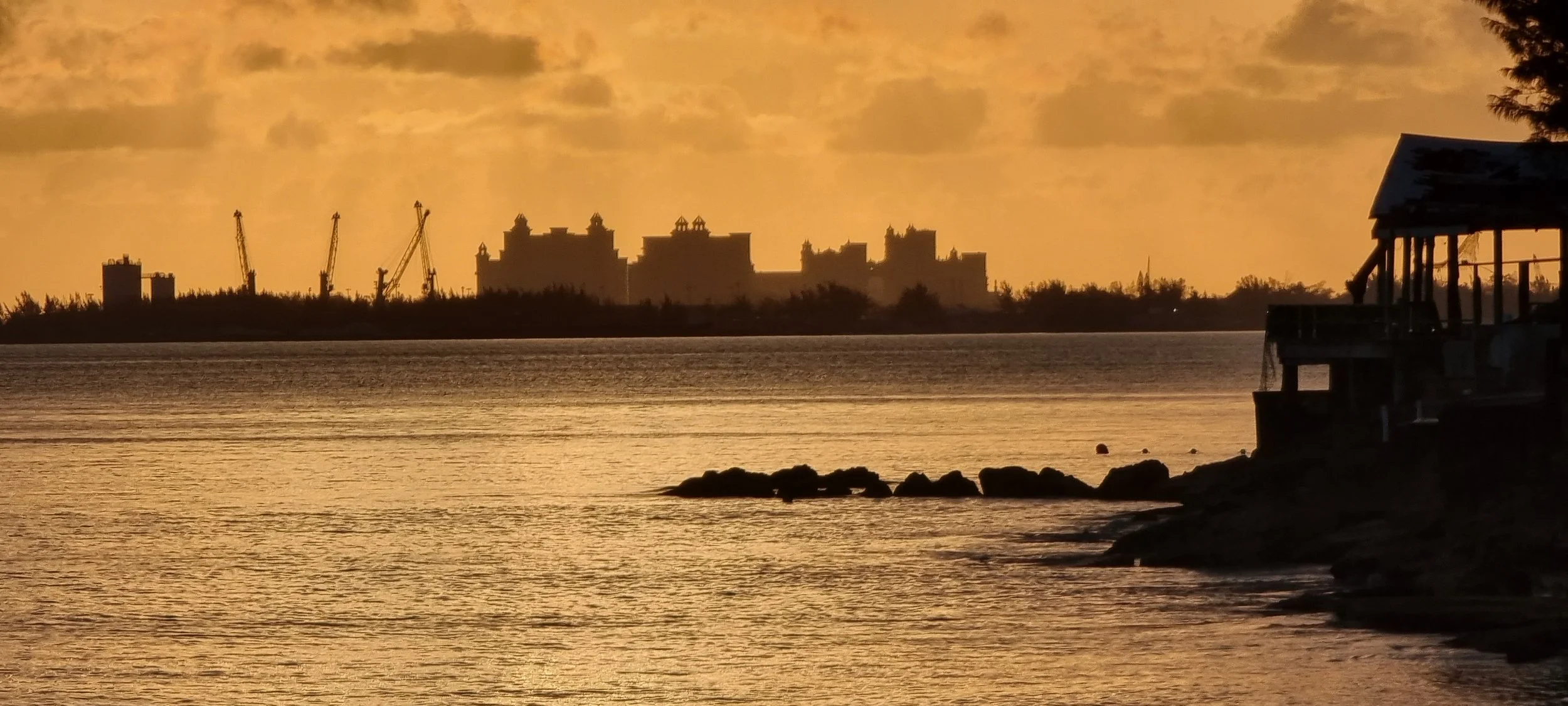 Sunset over a body of water with a silhouette of a house on the right and a distant industrial skyline with cranes in the background.