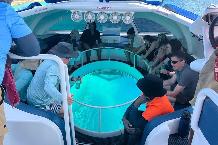 Group of people sitting around a circular hot tub on a boat, some with drinks, looking into the water, with a woman standing inside the hot tub, and a person in an orange shirt and black hat sitting on the side.