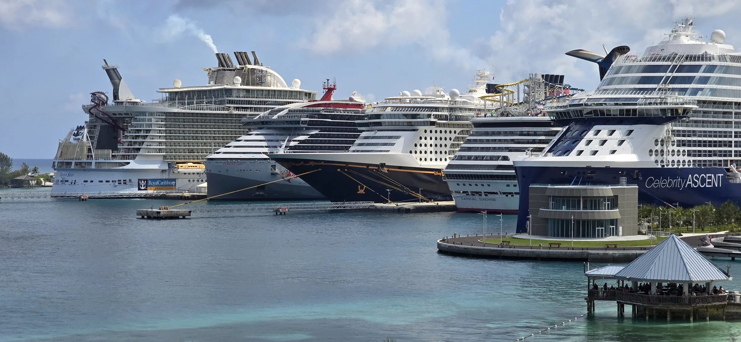 Multiple large cruise ships docked at a port, with water in the foreground and a small pier with a roofed pavilion on the right side.