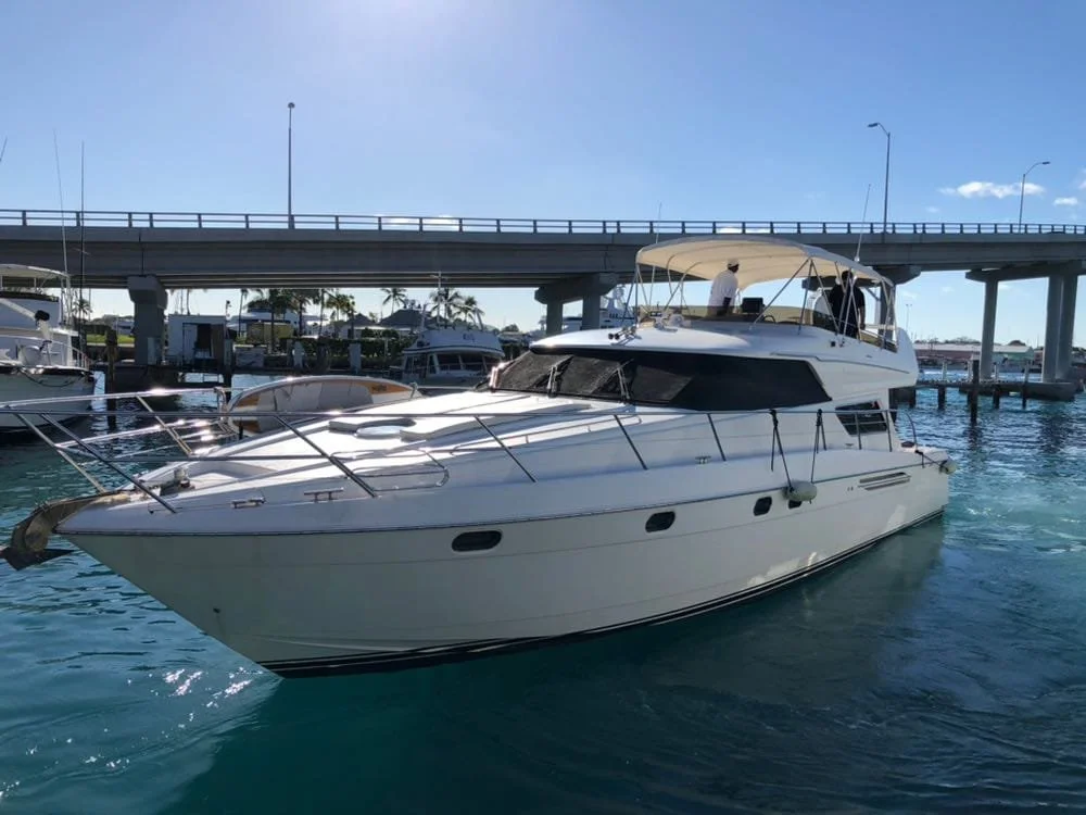 White motor yacht docked at marina under bridge, with clear blue sky and water.