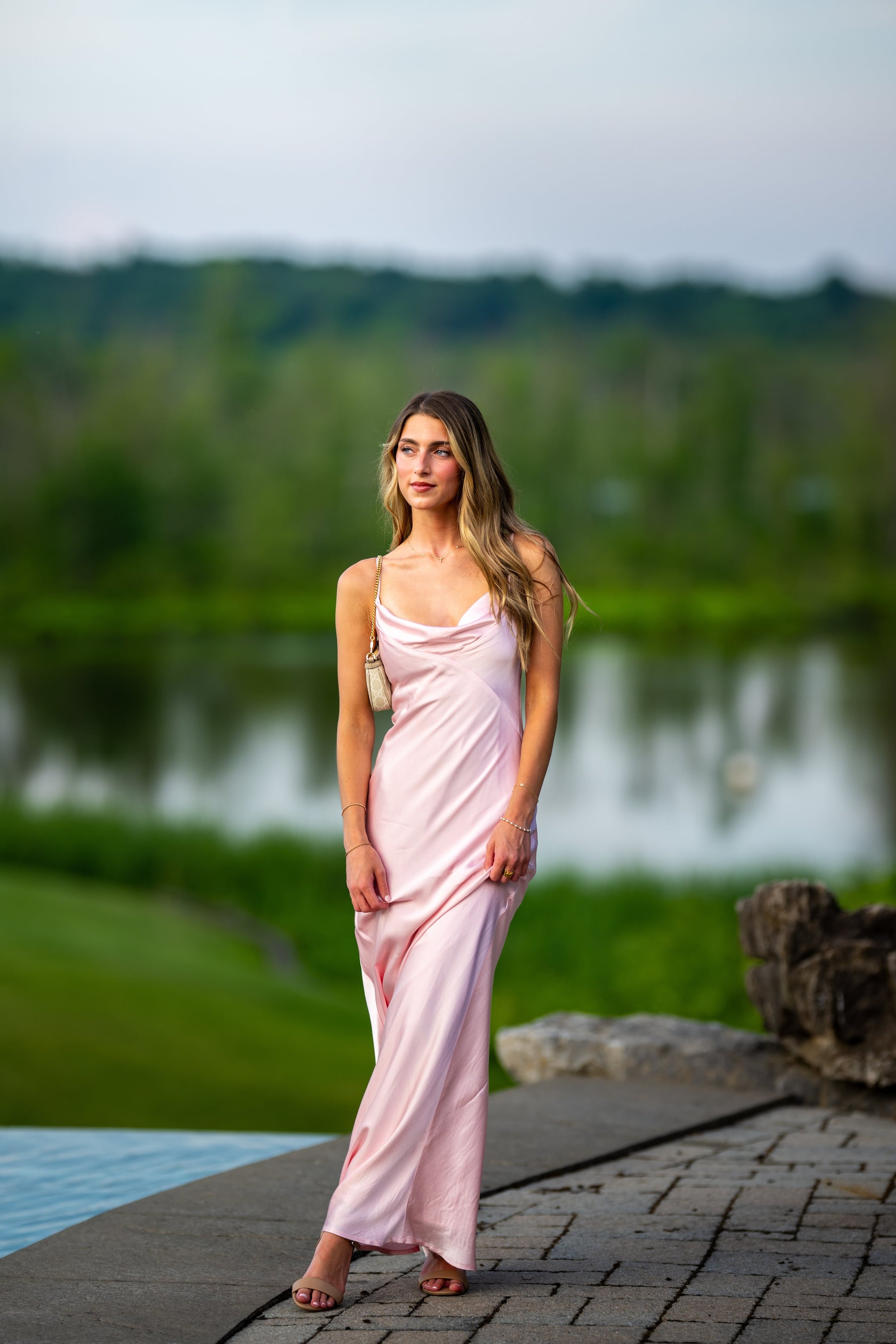 A woman in a light pink satin dress standing outdoors near a pond with greenery and trees in the background.
