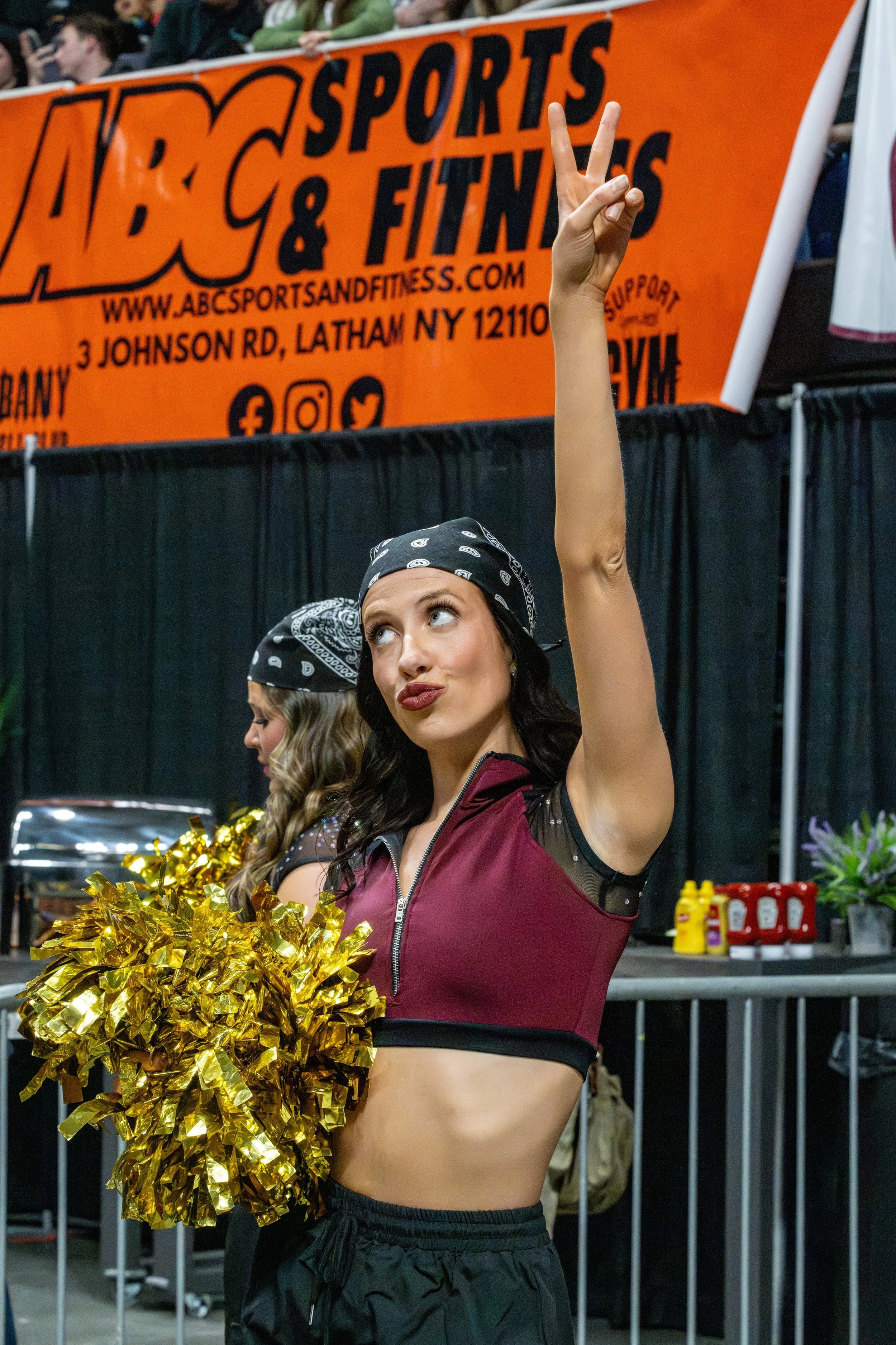 Cheerleader holding gold pom-poms with her arm raised making a peace sign in an indoor sports arena, with a black curtain and an orange banner advertising ABC Sports & Fitness in the background.