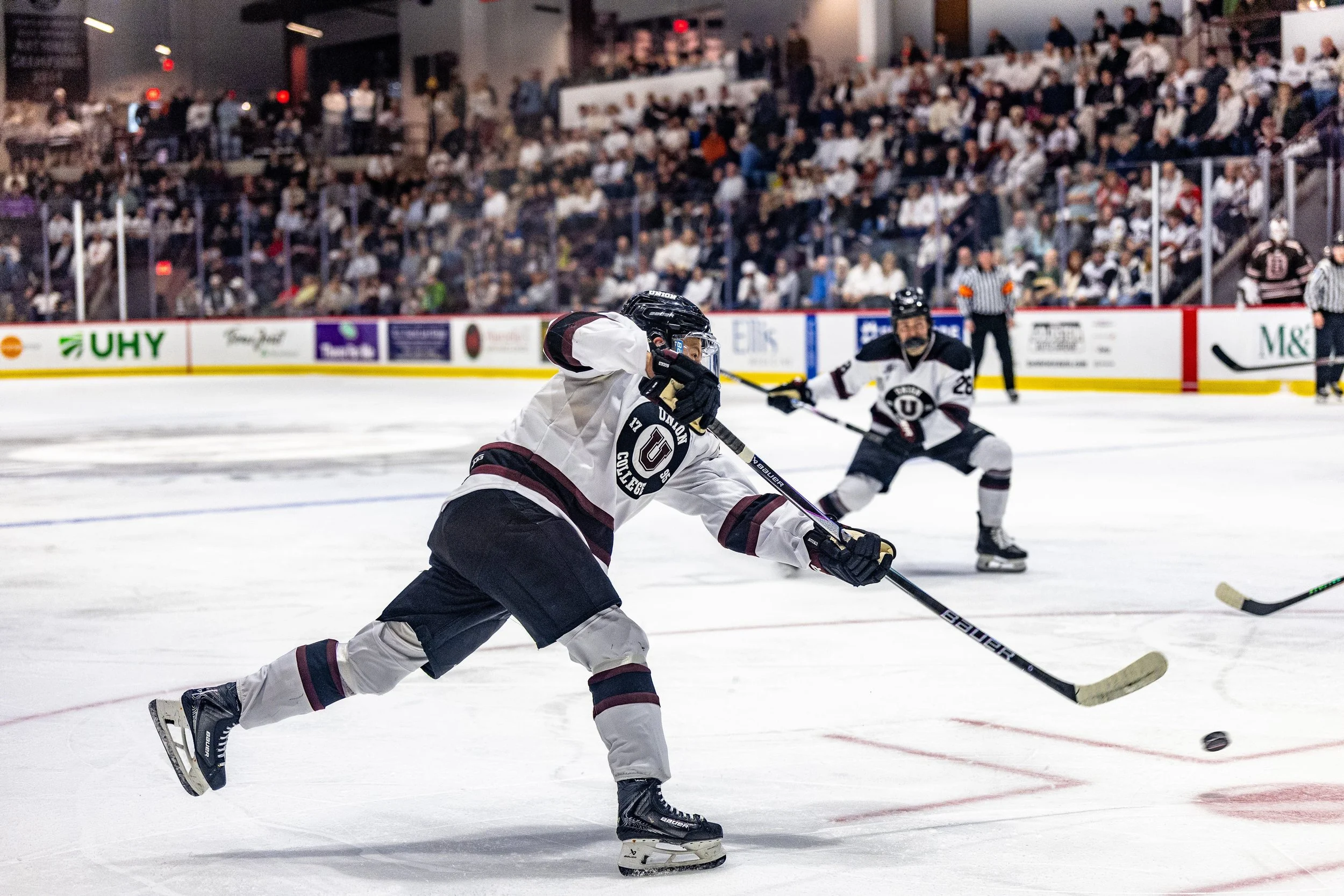 Ice hockey players in white jerseys playing on the rink, with one player in focus preparing to hit the puck, and a crowd of spectators watching from the stands.