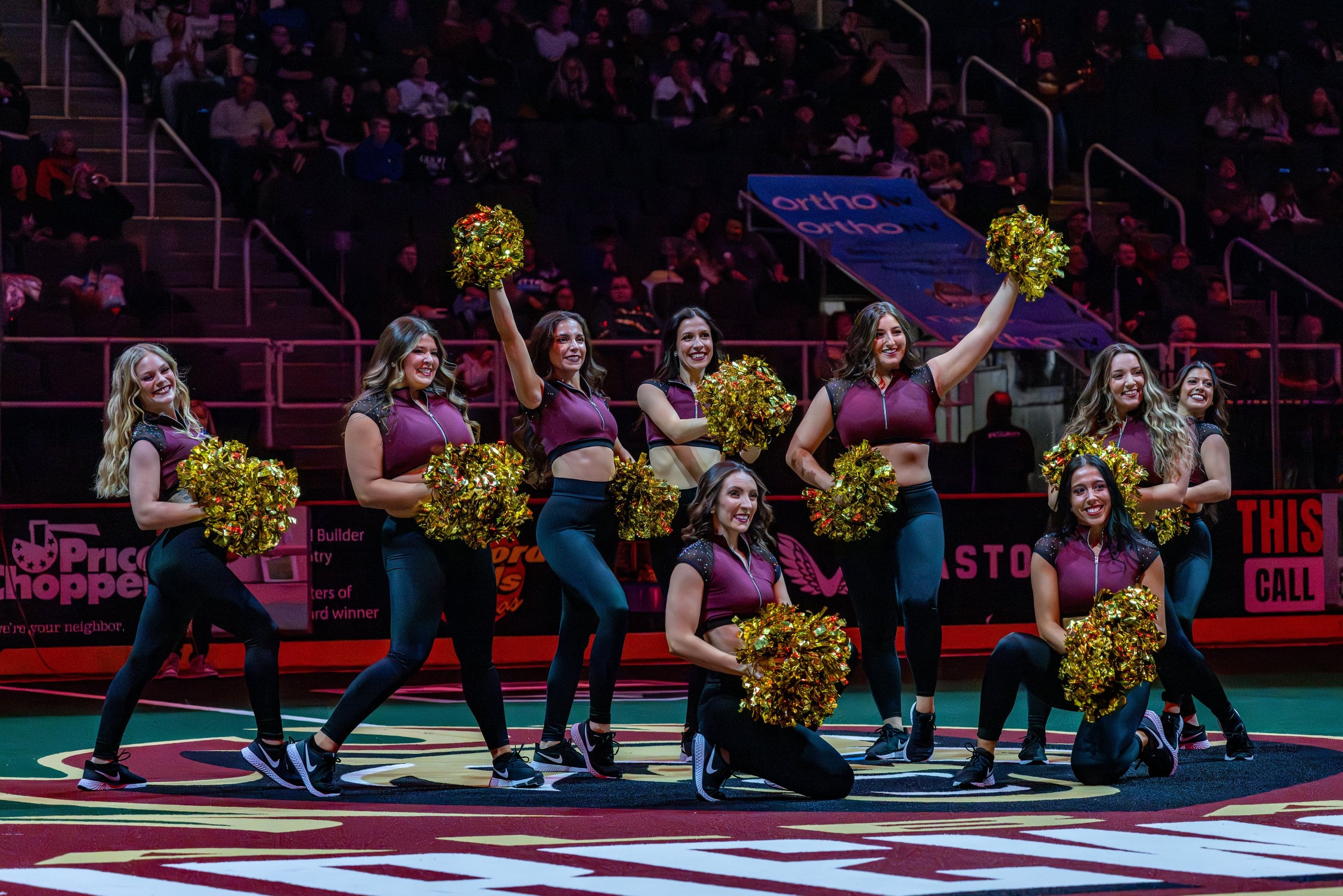Eight cheerleaders in matching maroon and black uniforms perform a routine on a sports court, holding gold pom-poms, with an audience in the background.