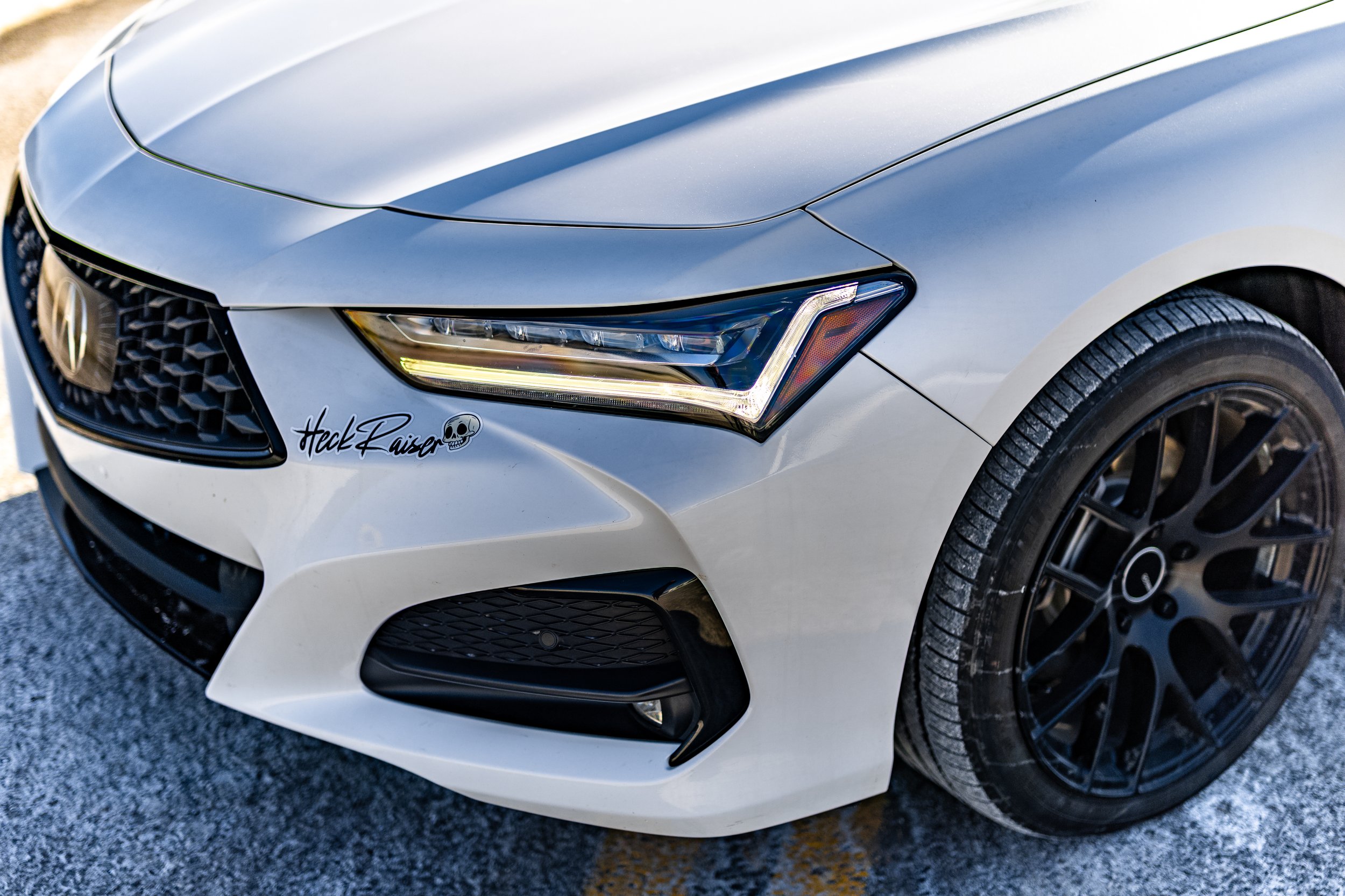 Close-up of the front right side of a white car with black wheels, a grille, and a headlight, parked on a paved surface with snow.