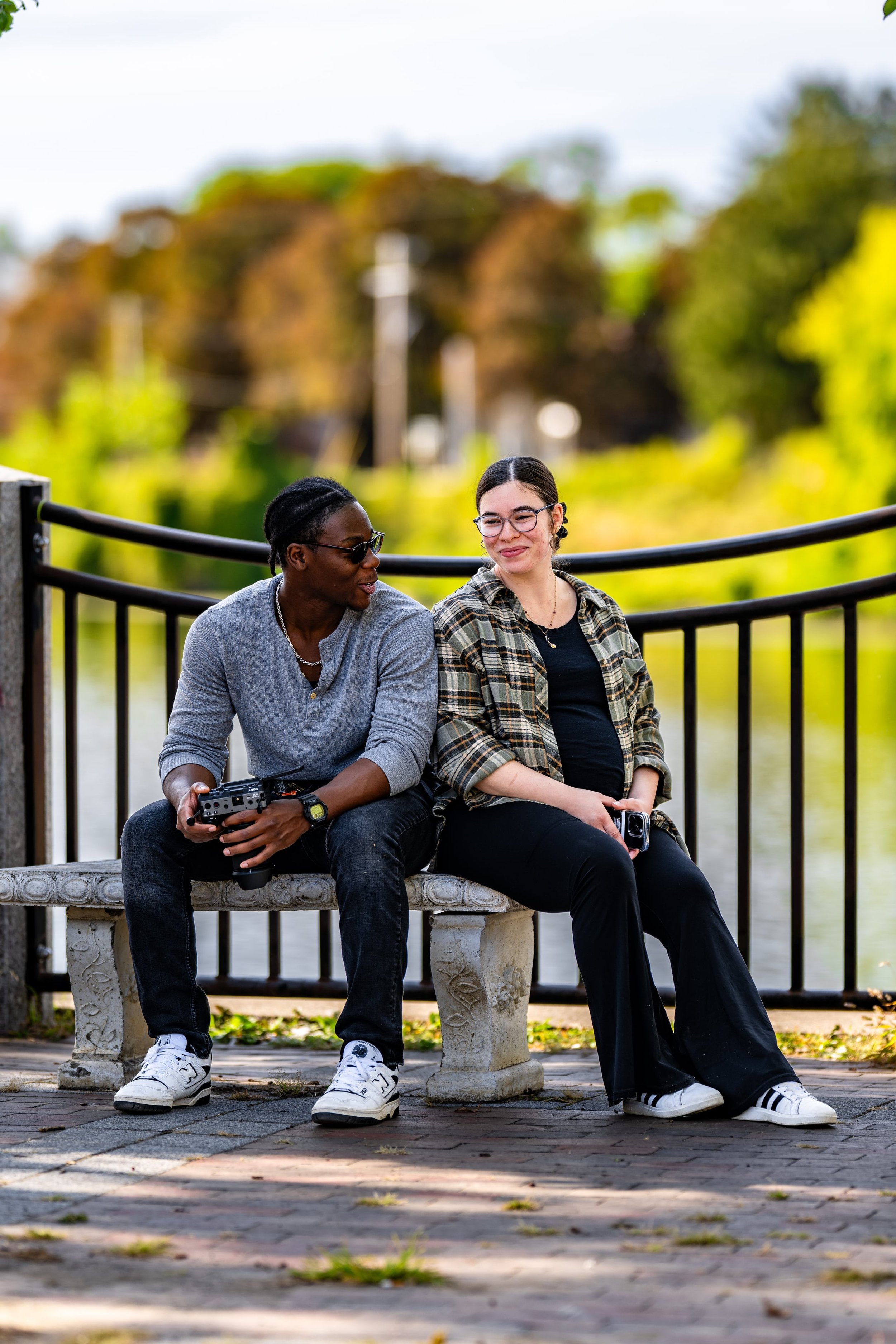 Two friends sitting on a park bench near a body of water, having a conversation with trees and sky in the background.