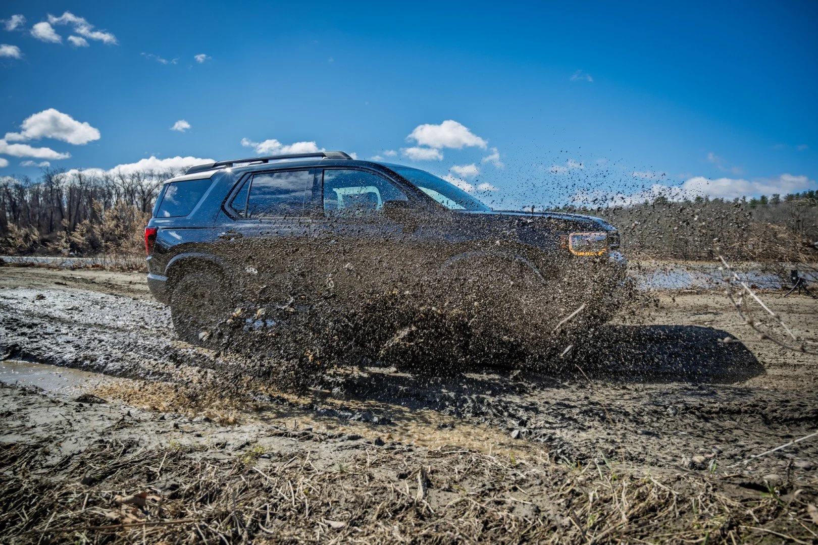 A black SUV driving through a muddy, dirt terrain, splashing mud and water, with a blue sky and scattered clouds above.