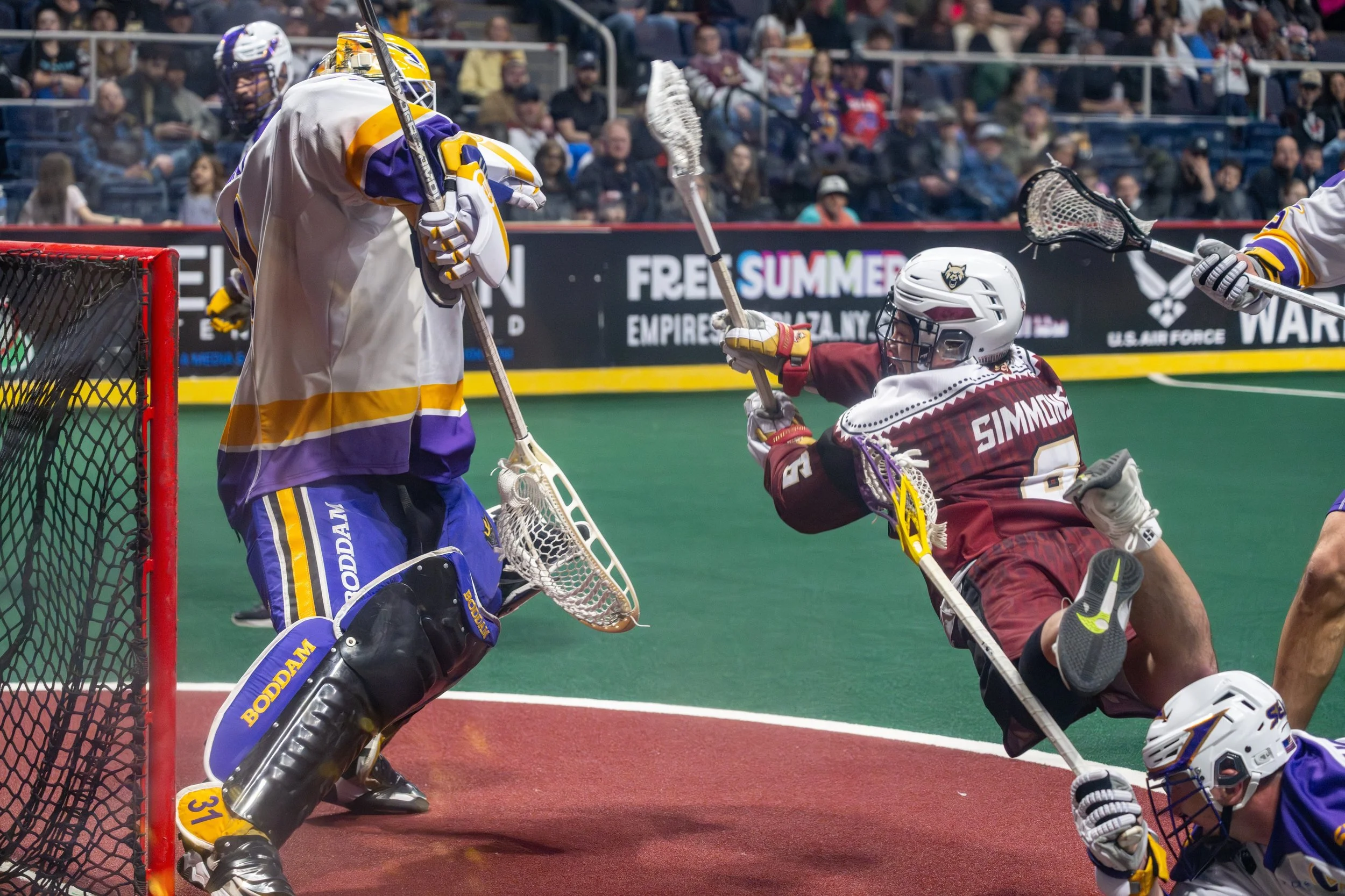 Lacrosse players battling for the ball near the goal, with one player falling on the ground and another goalie in position ready to defend.
