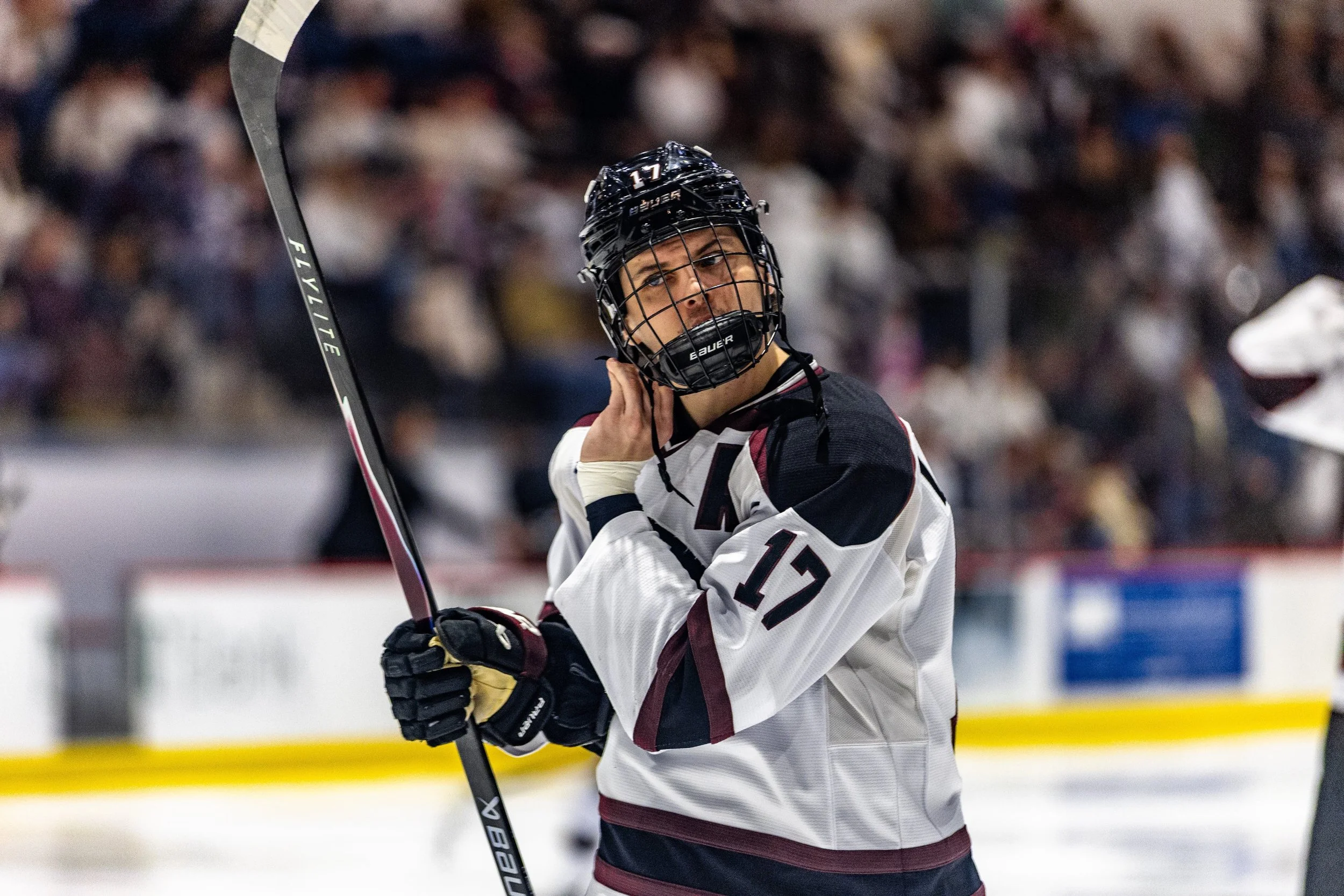 An ice hockey player in a white jersey with black and maroon accents, wearing a black helmet, holding a hockey stick, and touching his neck during a game, with a blurred crowd in the background.