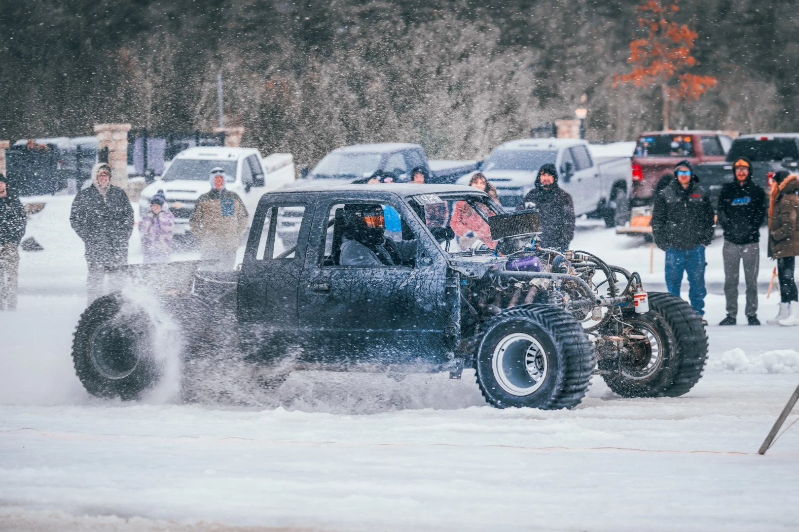 A snow-covered area with a vehicle designed for racing or testing, with large tires and an exposed engine, moving quickly and creating splashes of snow behind it. Several people are watching in the background, with parked cars and trees visible.