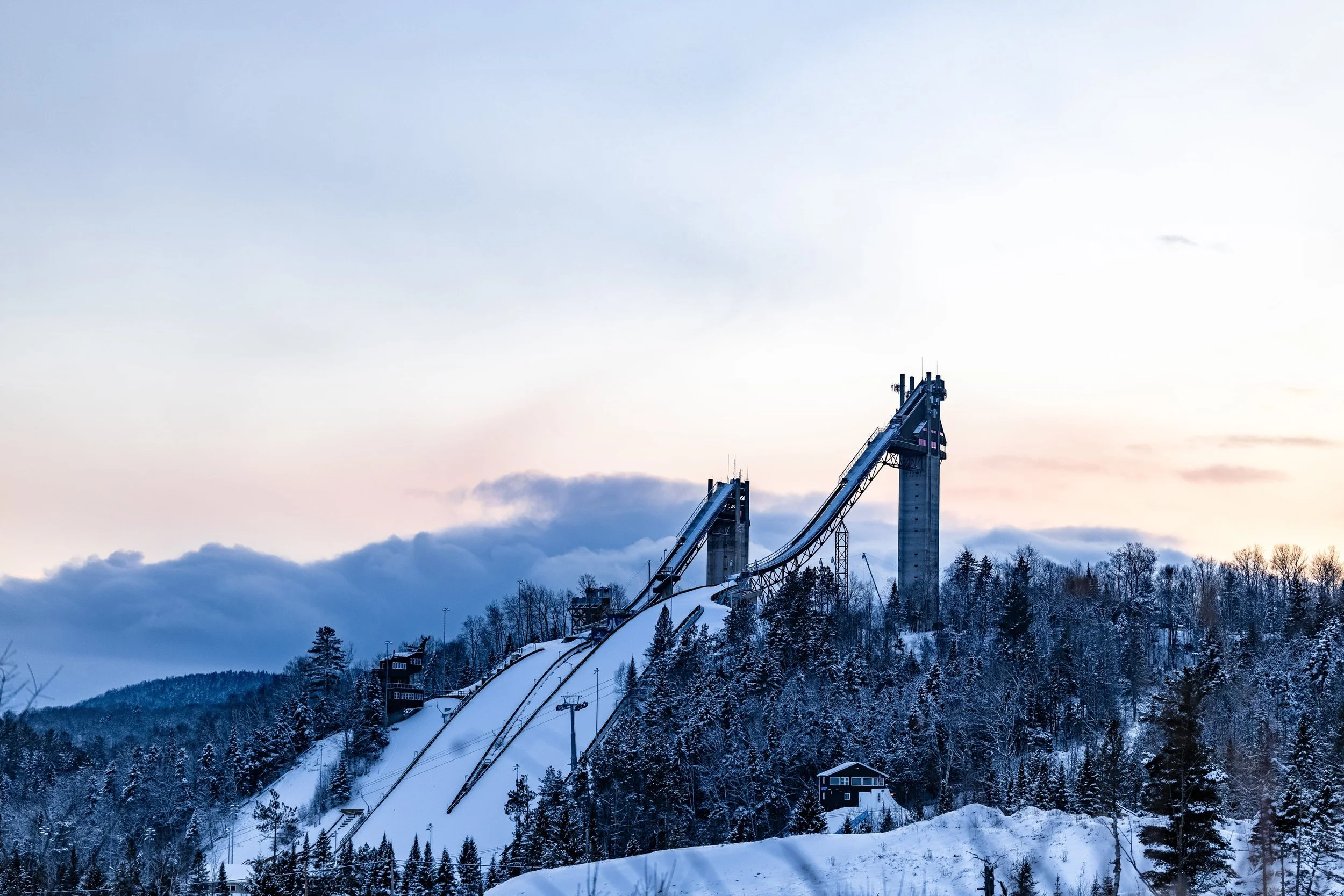 Ski jump ramp on a snowy mountain, surrounded by trees, with a cloudy sky at dusk.