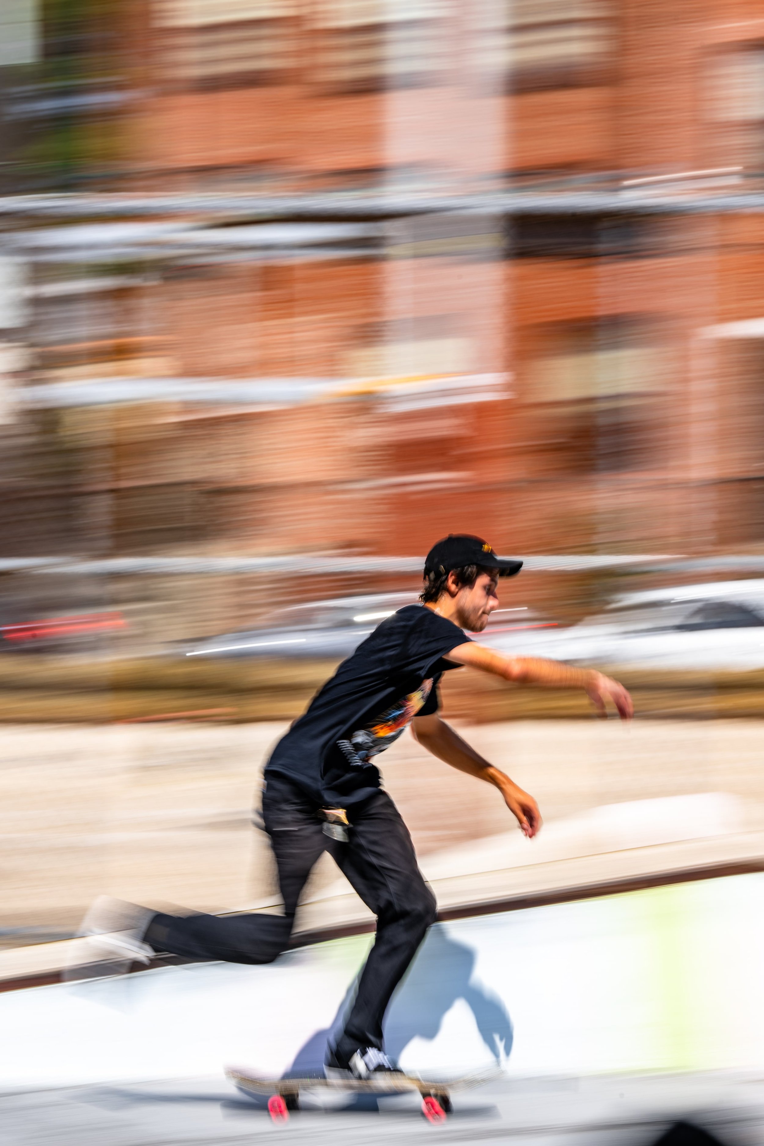 A young man skateboarding outdoors with a blurred background of buildings and parked cars, indicating motion and speed.