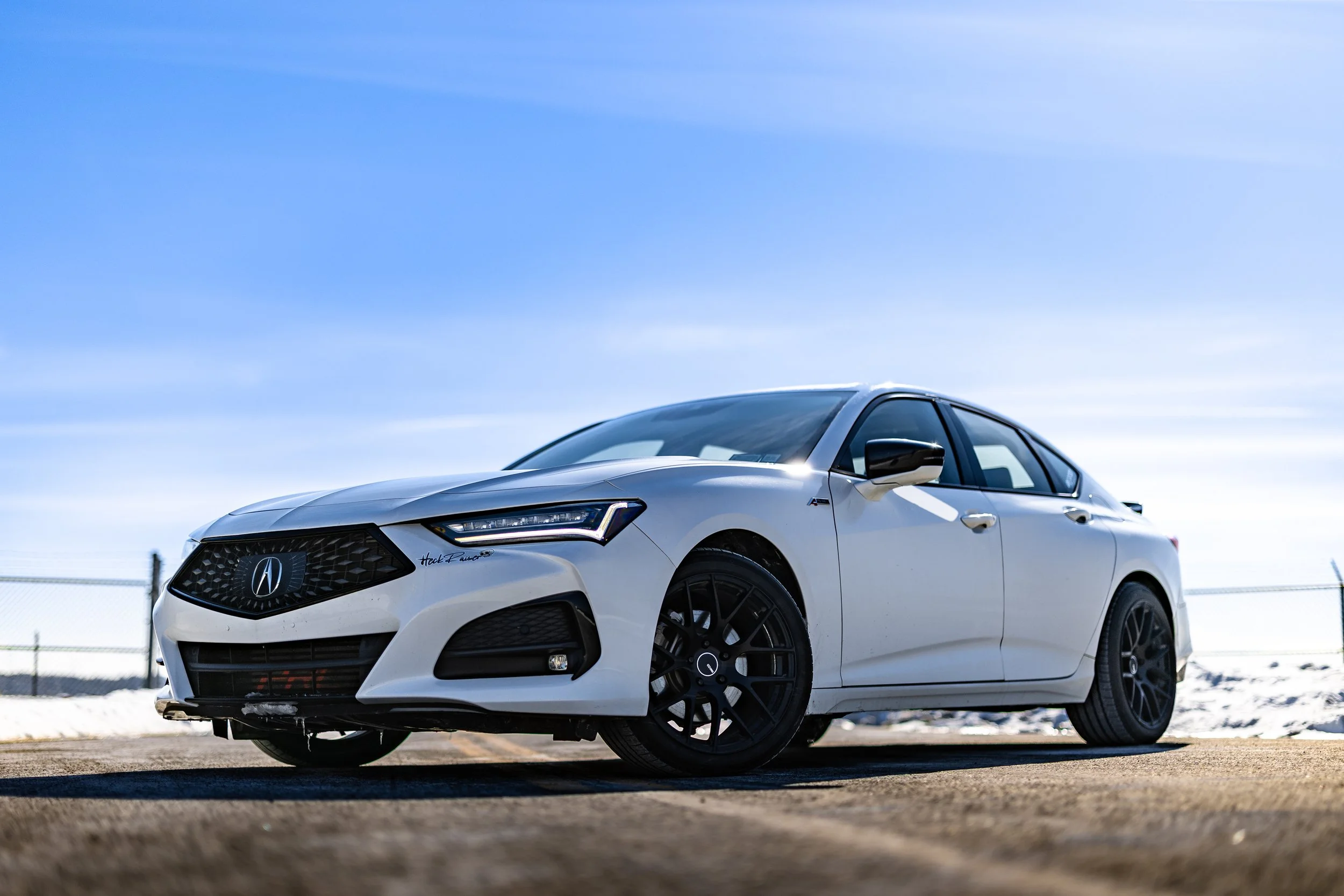 A white Acura sedan parked outdoors on a dirt surface with a chain-link fence and snow in the background under a clear blue sky.
