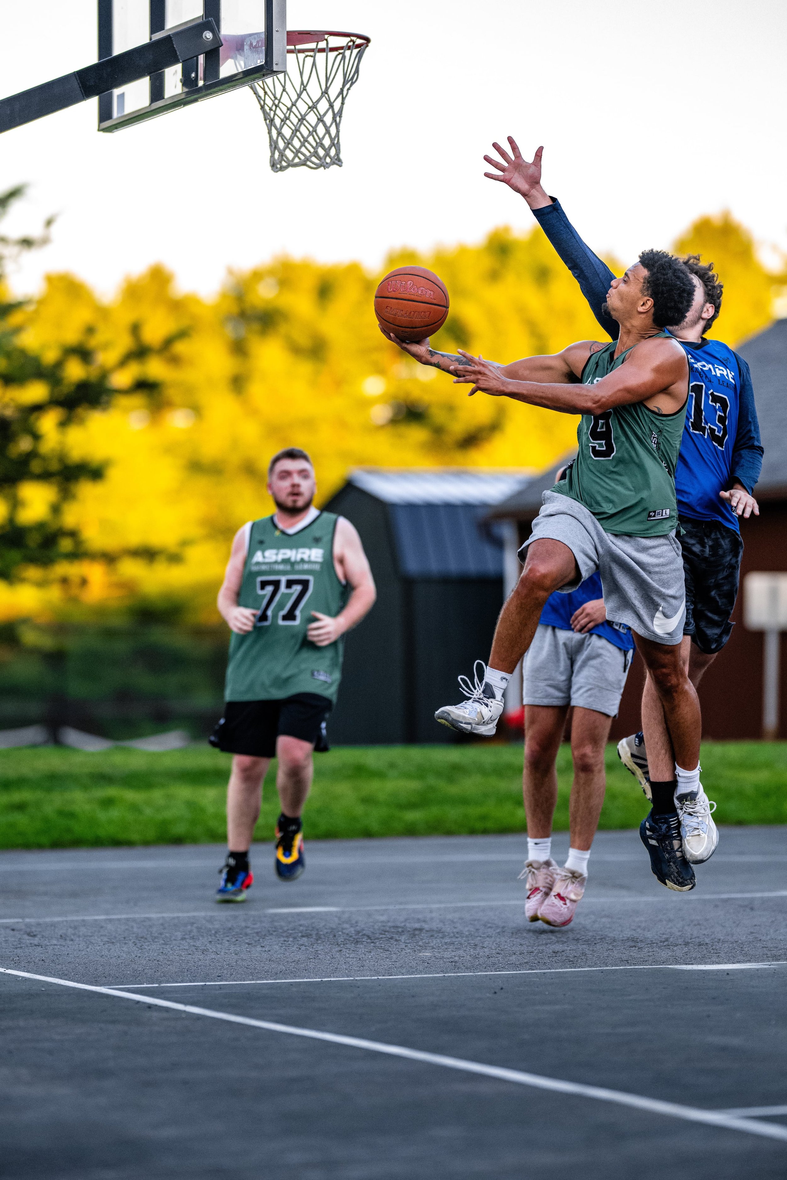 People playing basketball outdoors during the daytime, with one player attempting a shot while others are around him on the court.