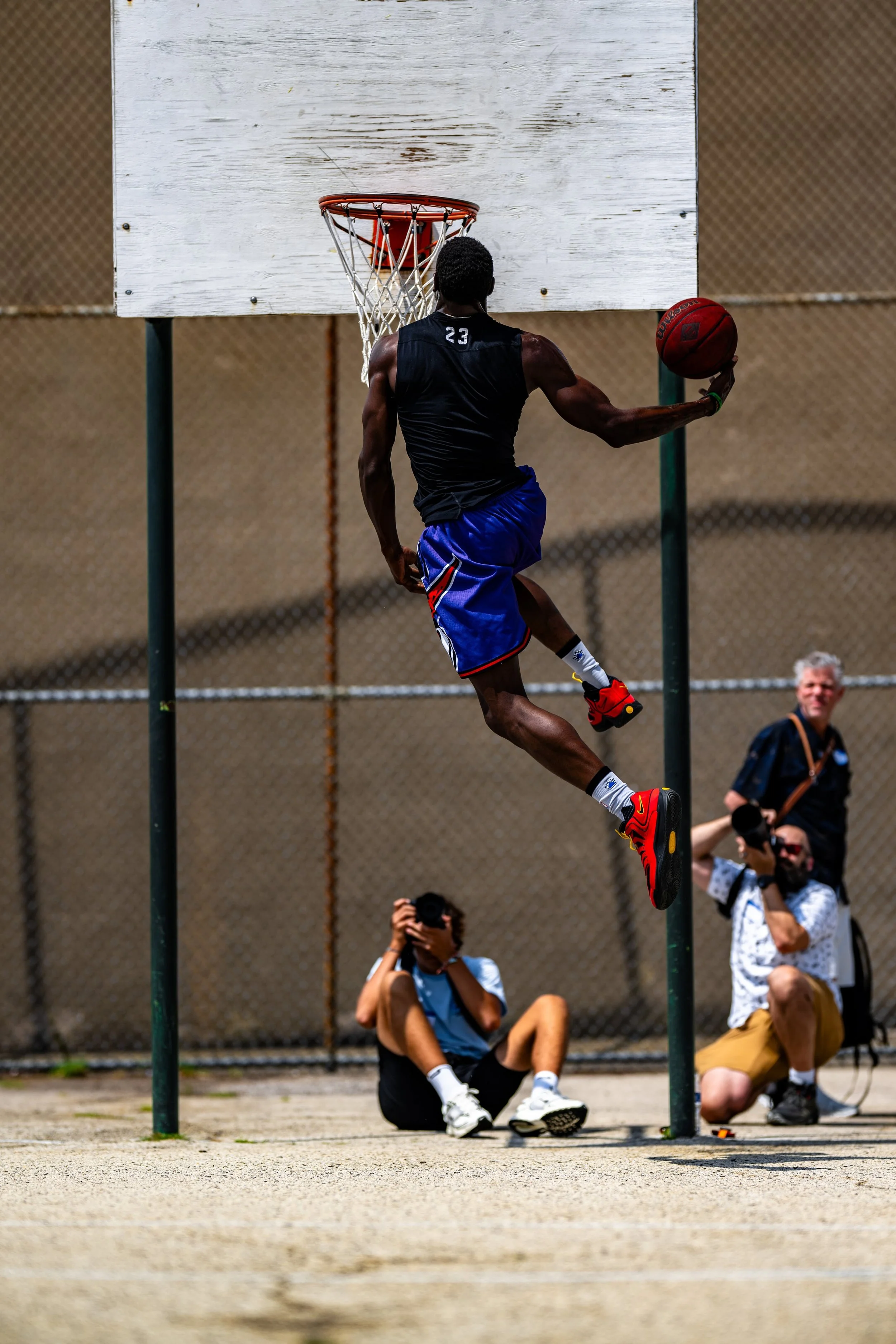 A man wearing a black sleeveless shirt and blue shorts is mid-air, dunking a basketball in an outdoor court. Three photographers are in the background, one sitting on the ground and two standing, taking pictures of him.