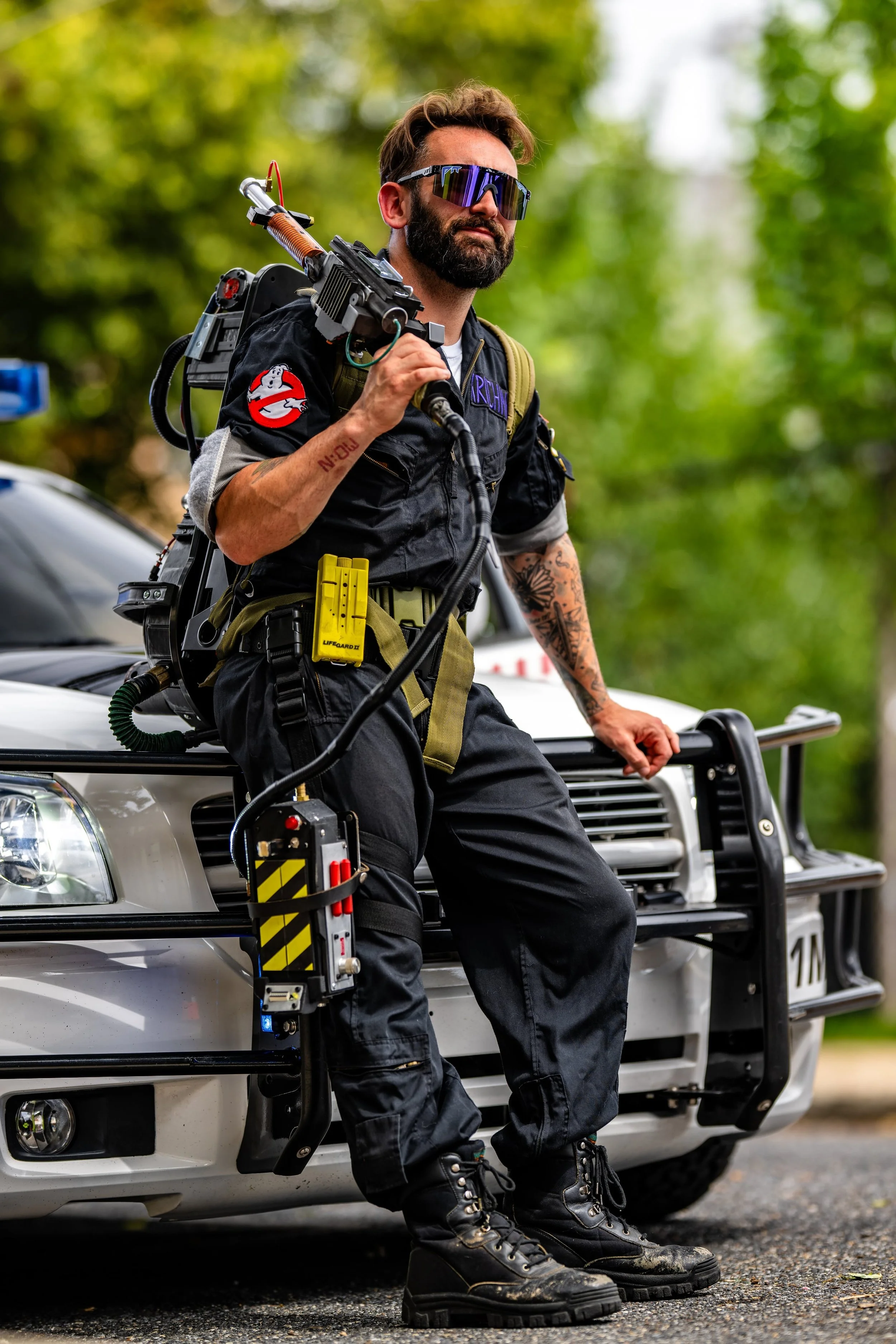 A man dressed as a Ghostbuster, holding a proton pack weapon, sitting on the hood of a police car with a green wooded background.