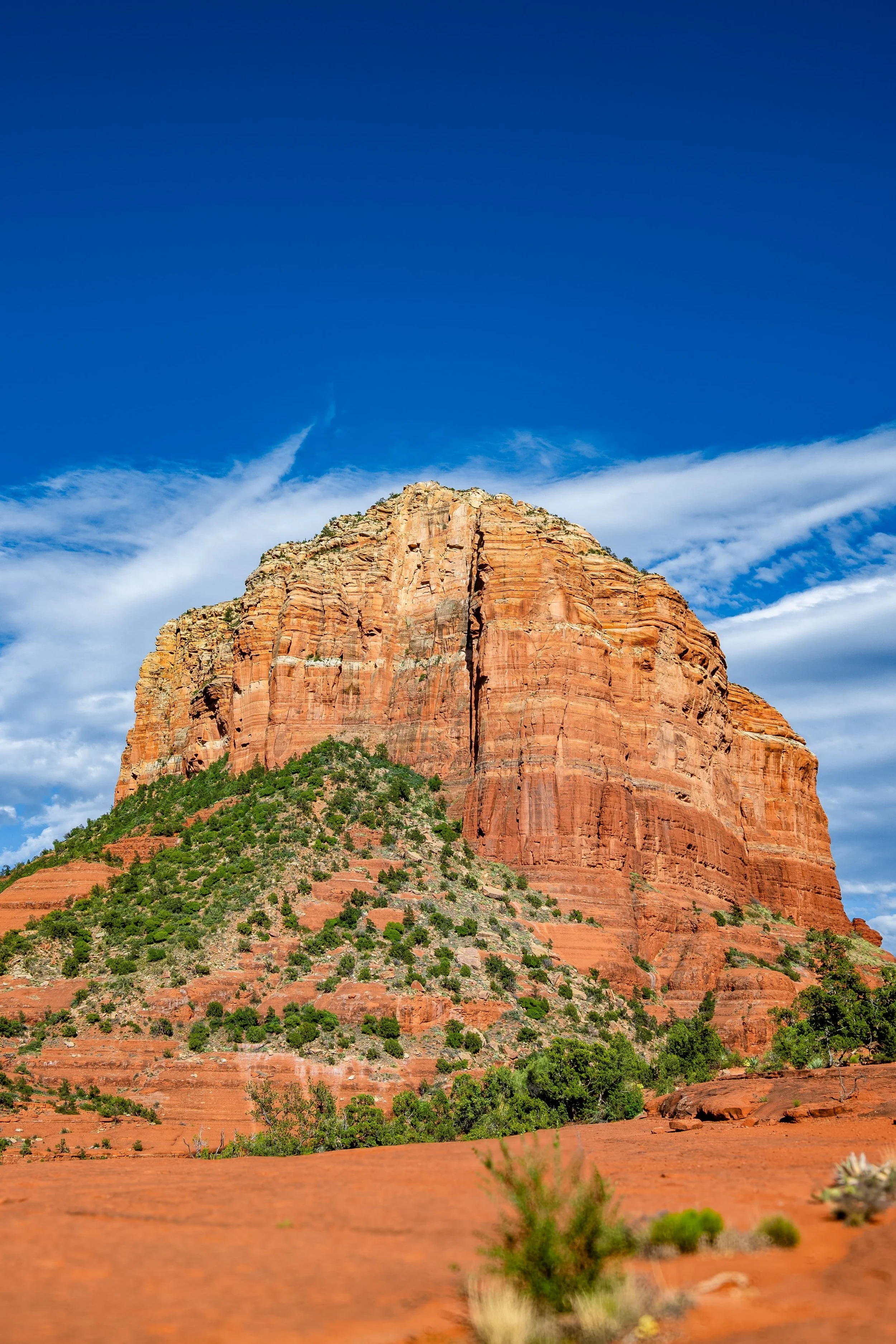 A large red sandstone cliff against a partly cloudy blue sky with some green vegetation at its base.