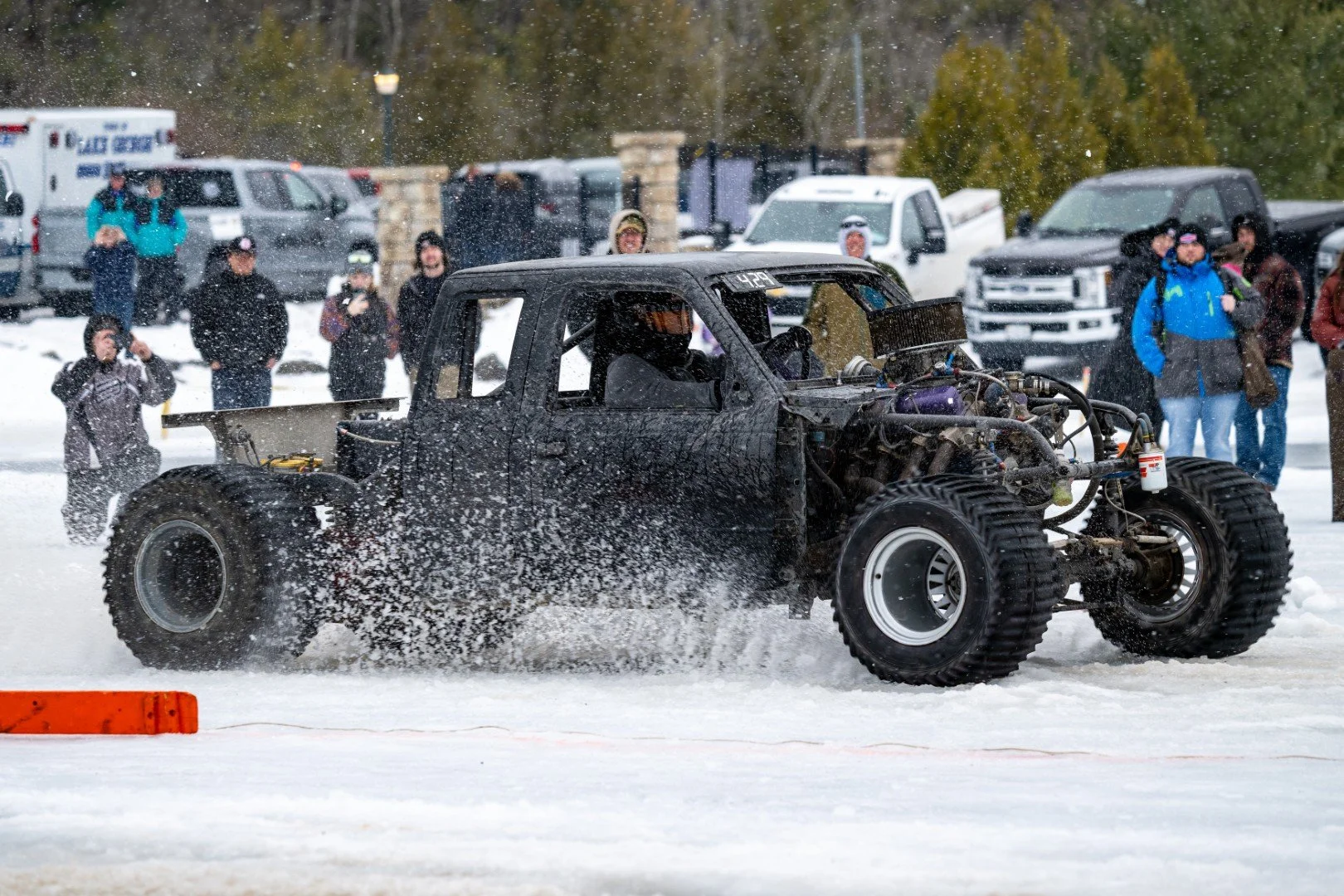 An off-road vehicle racing through snow, kicking up a spray of snow behind it, with spectators watching in the background.