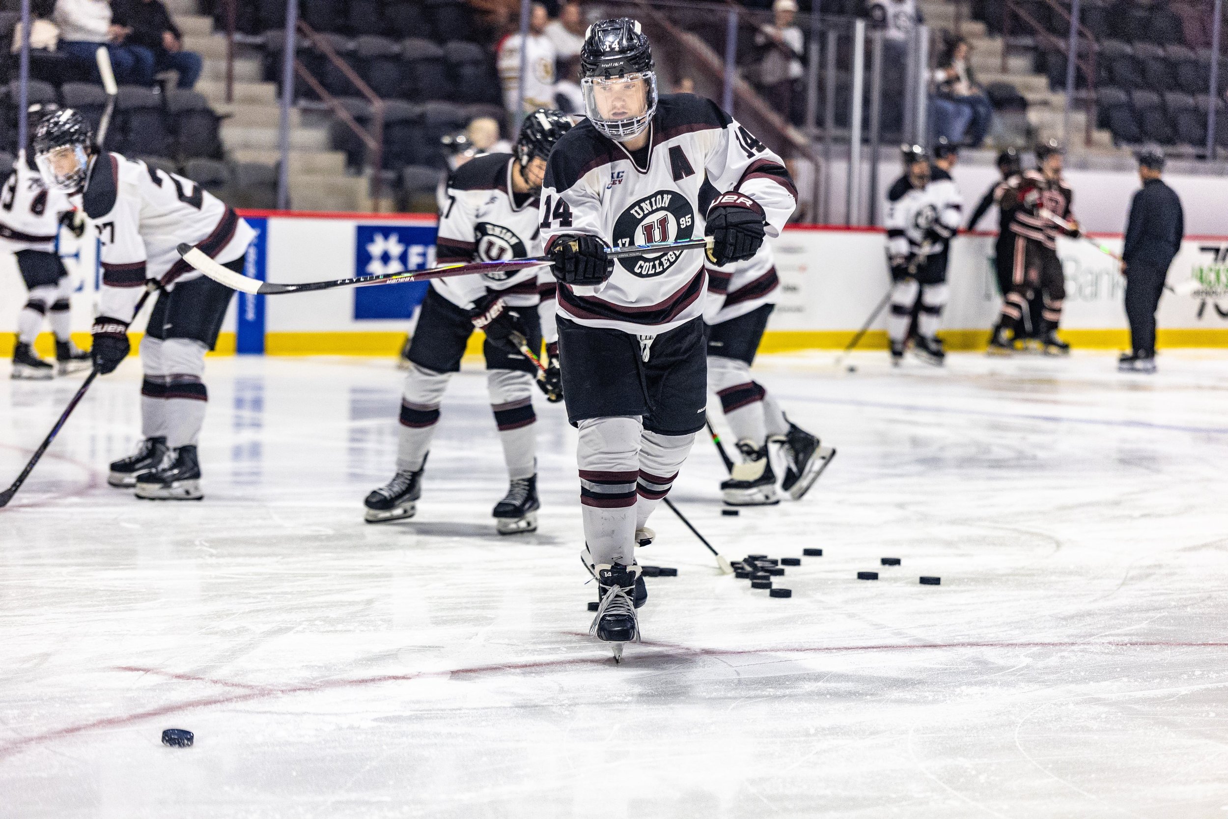Hockey players in white jerseys practice on the ice rink, with one player in the foreground handling puck training equipment.