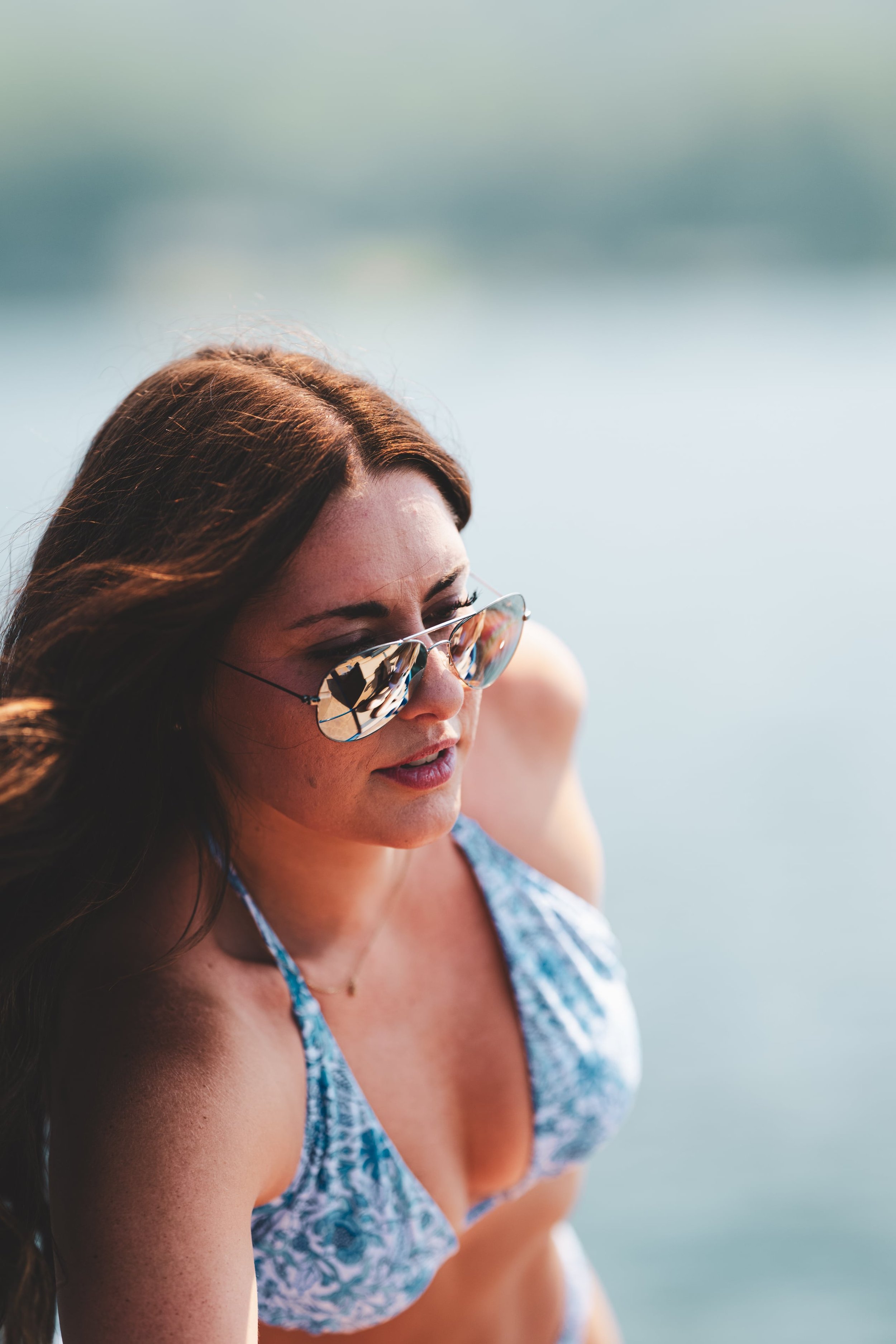 Woman with long brown hair wearing sunglasses and a blue and white patterned bikini at the beach.