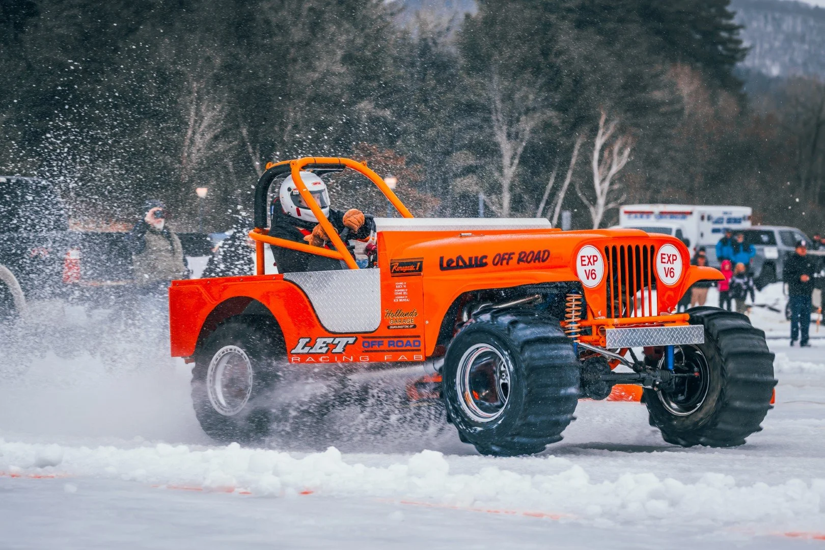 An orange off-road racing vehicle driving through snow, kicking up snow spray behind it, with spectators and trees in the background.