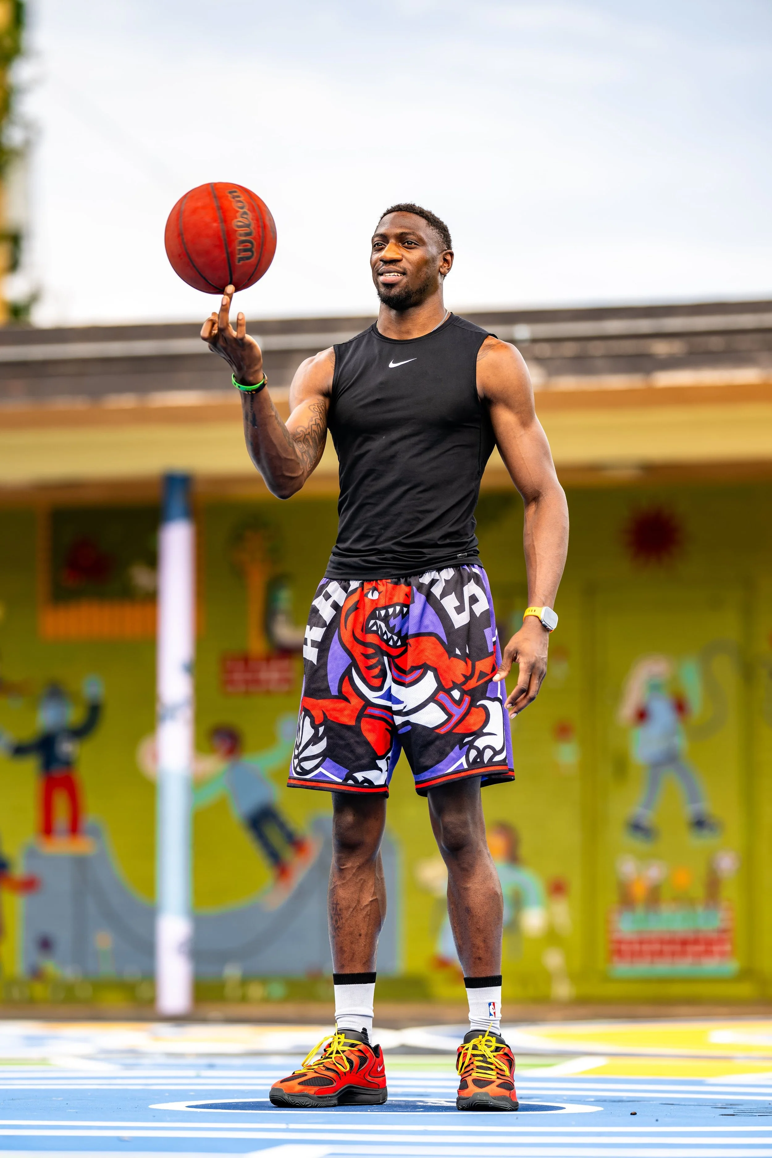 A man in athletic clothing, including a black tank top and colorful shorts with a dragon design, spinning a basketball on his finger on an outdoor court.