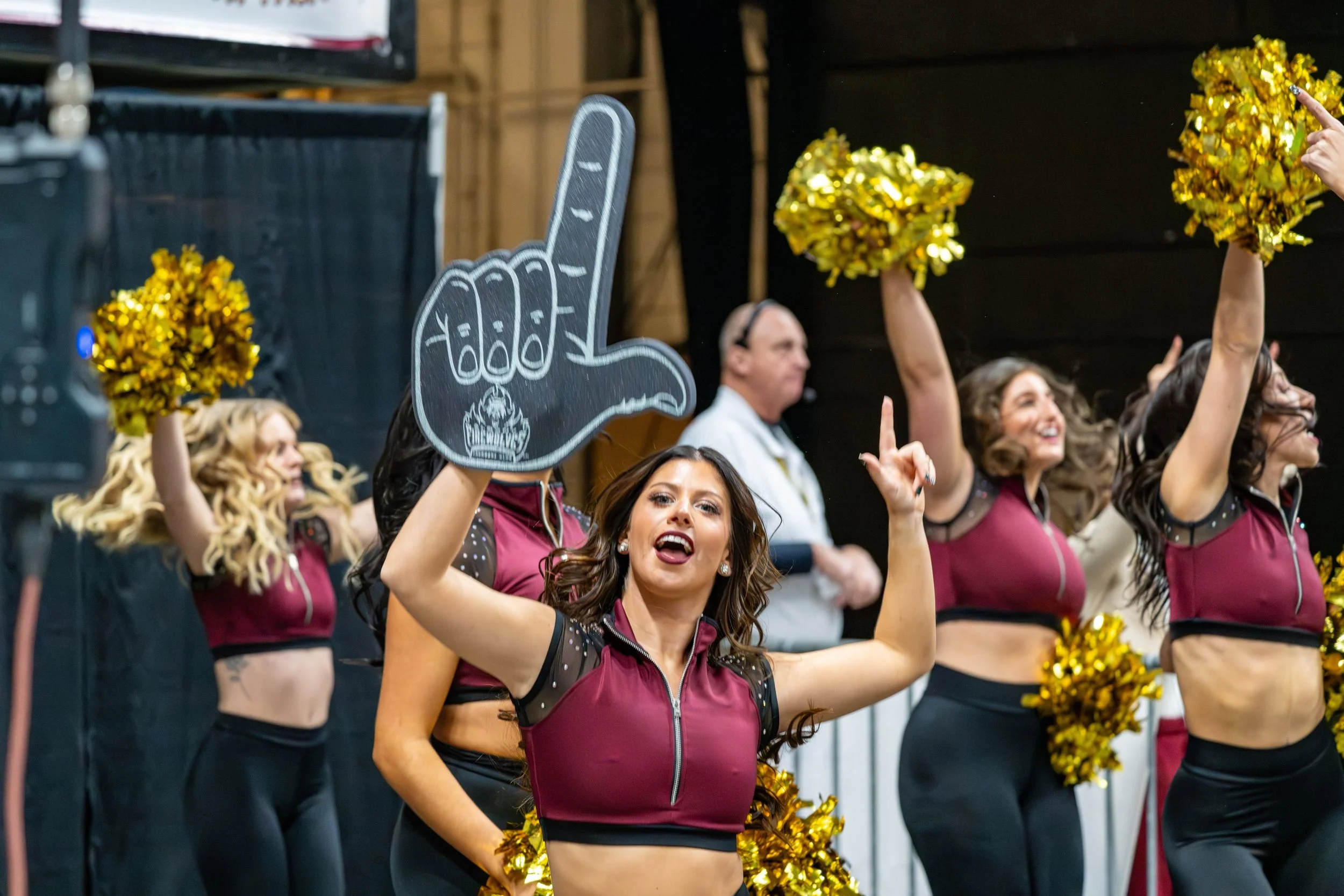 Cheerleaders performing with gold pom-poms, one cheerleader holding a chalkboard cutout of a hand with a cupcake drawing, in a cheerleading routine.