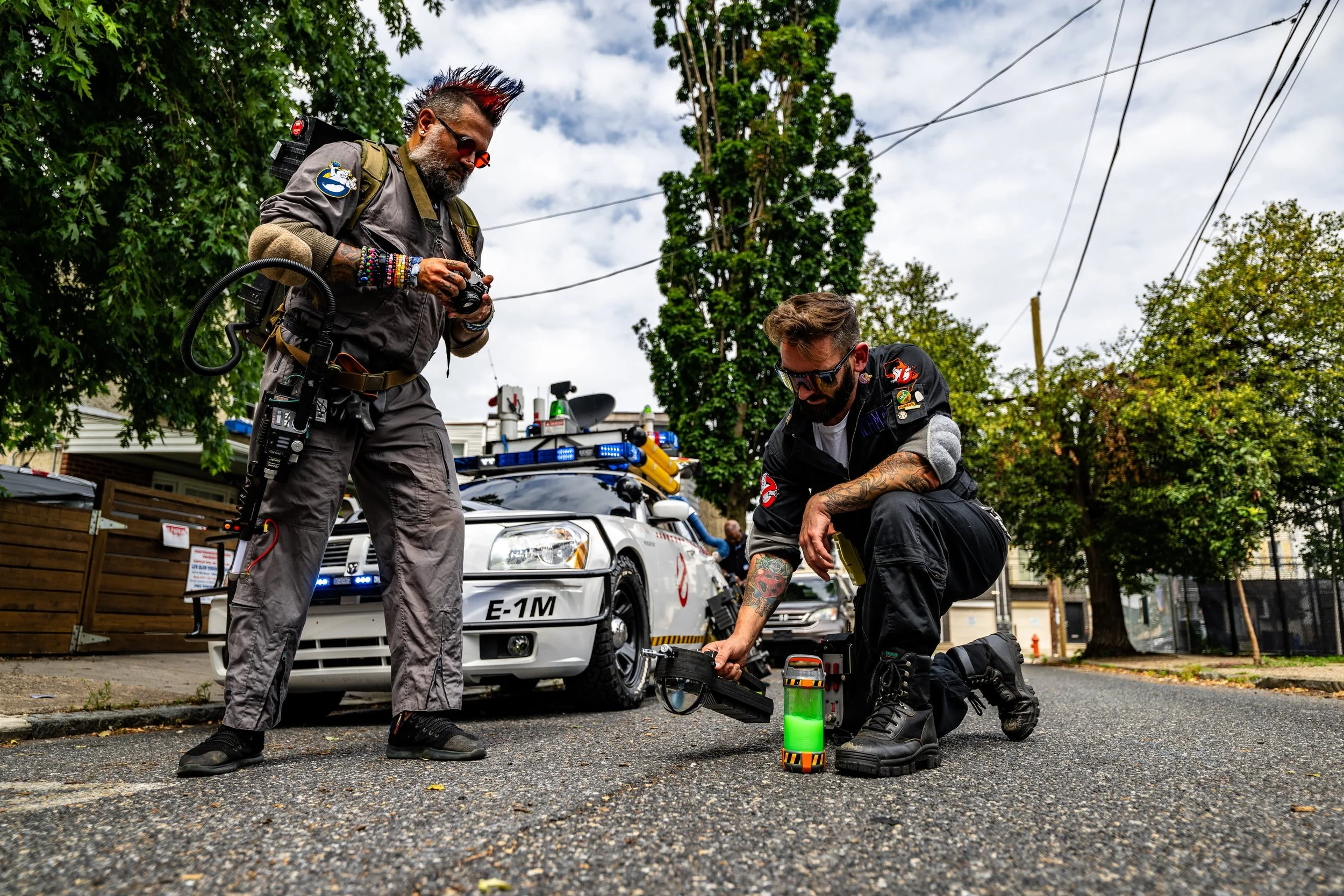 Two men dressed as Ghostbusters inspecting a proton pack on a street with a Ghostbusters-themed police car and trees in the background.