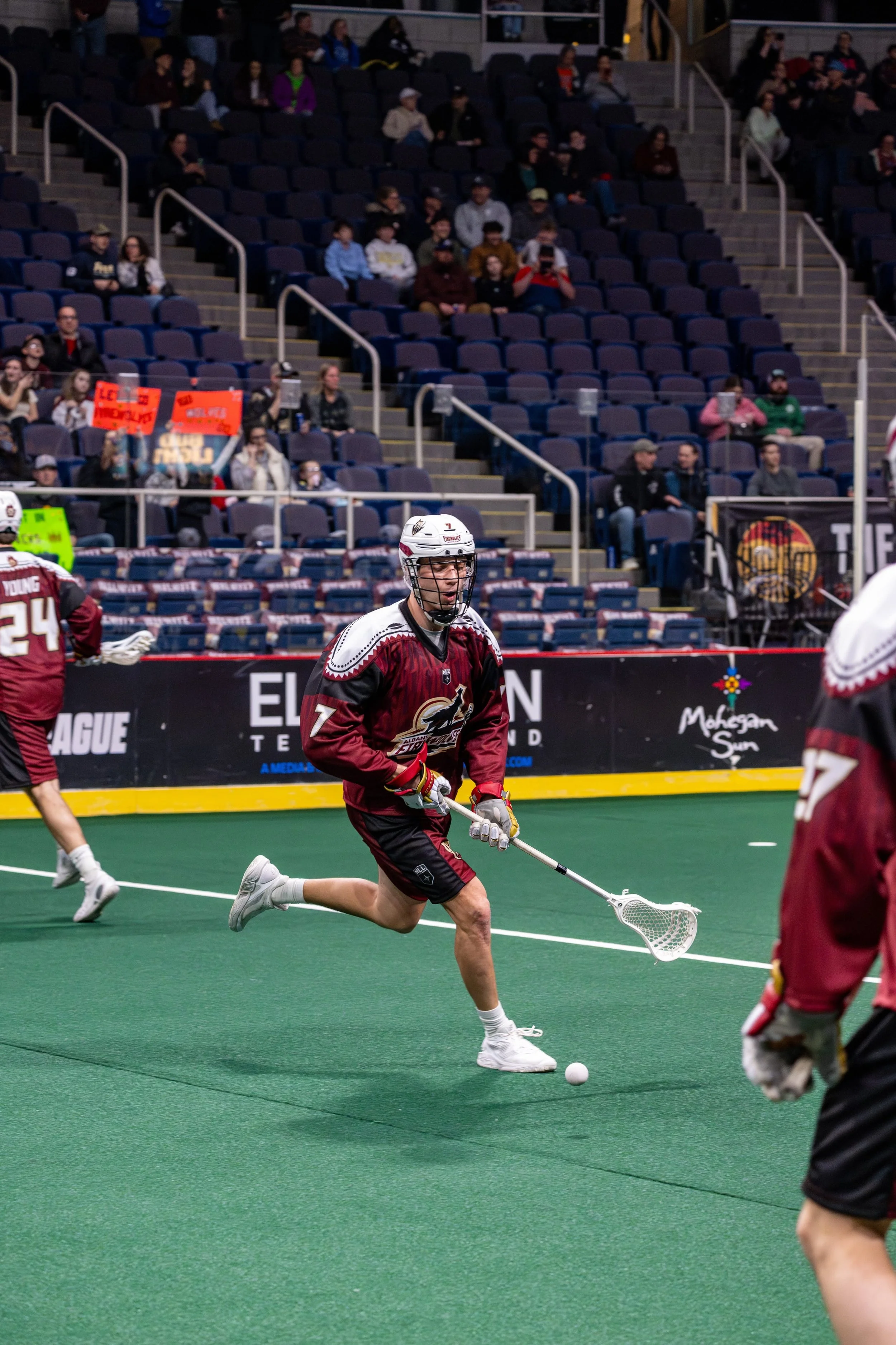 Indoor lacrosse game with players in maroon and black jerseys, one player running and reaching for a ball on green turf, spectators seated in the background.