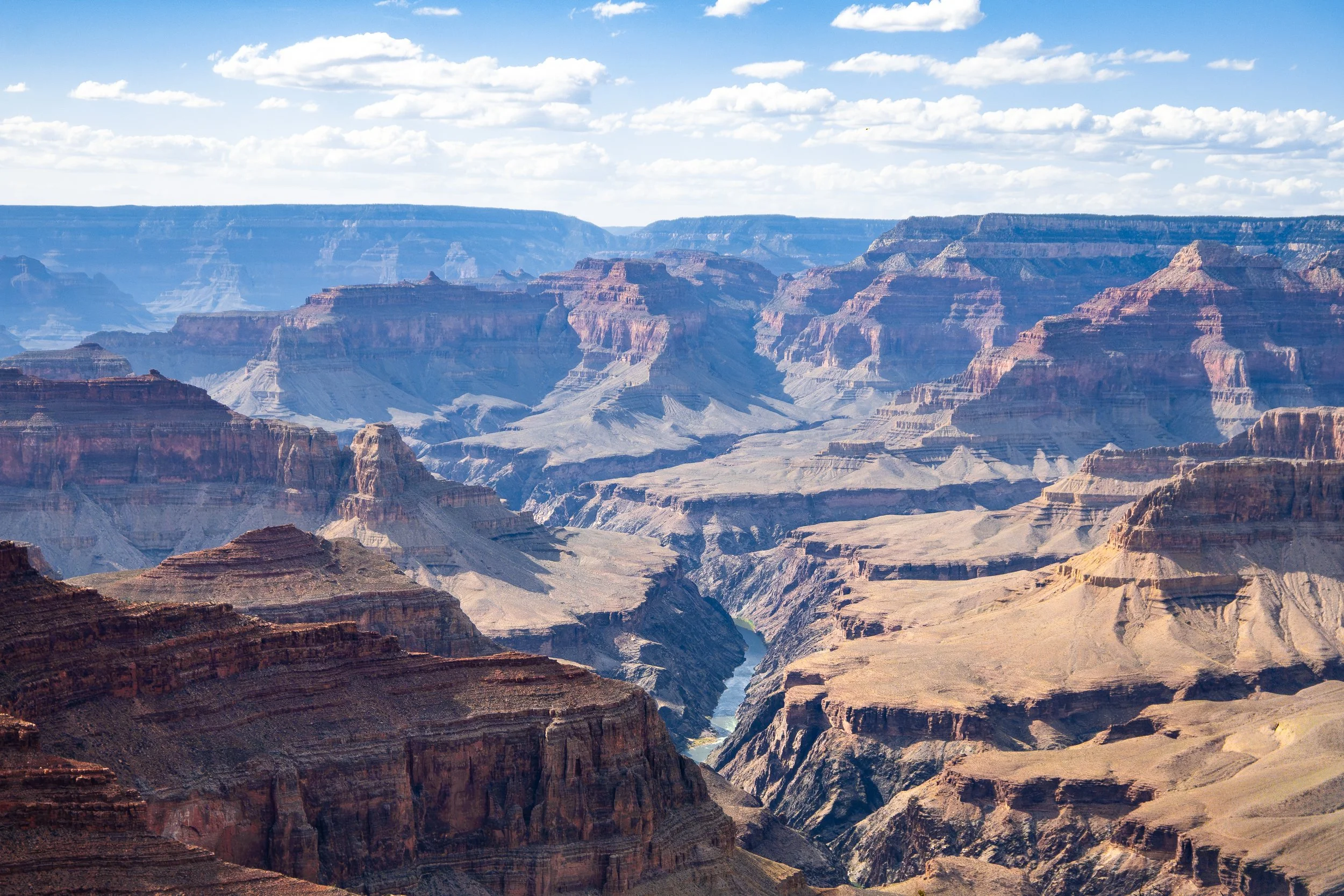 Grand Canyon landscape with layered rock formations and a river at the bottom under a partly cloudy sky.