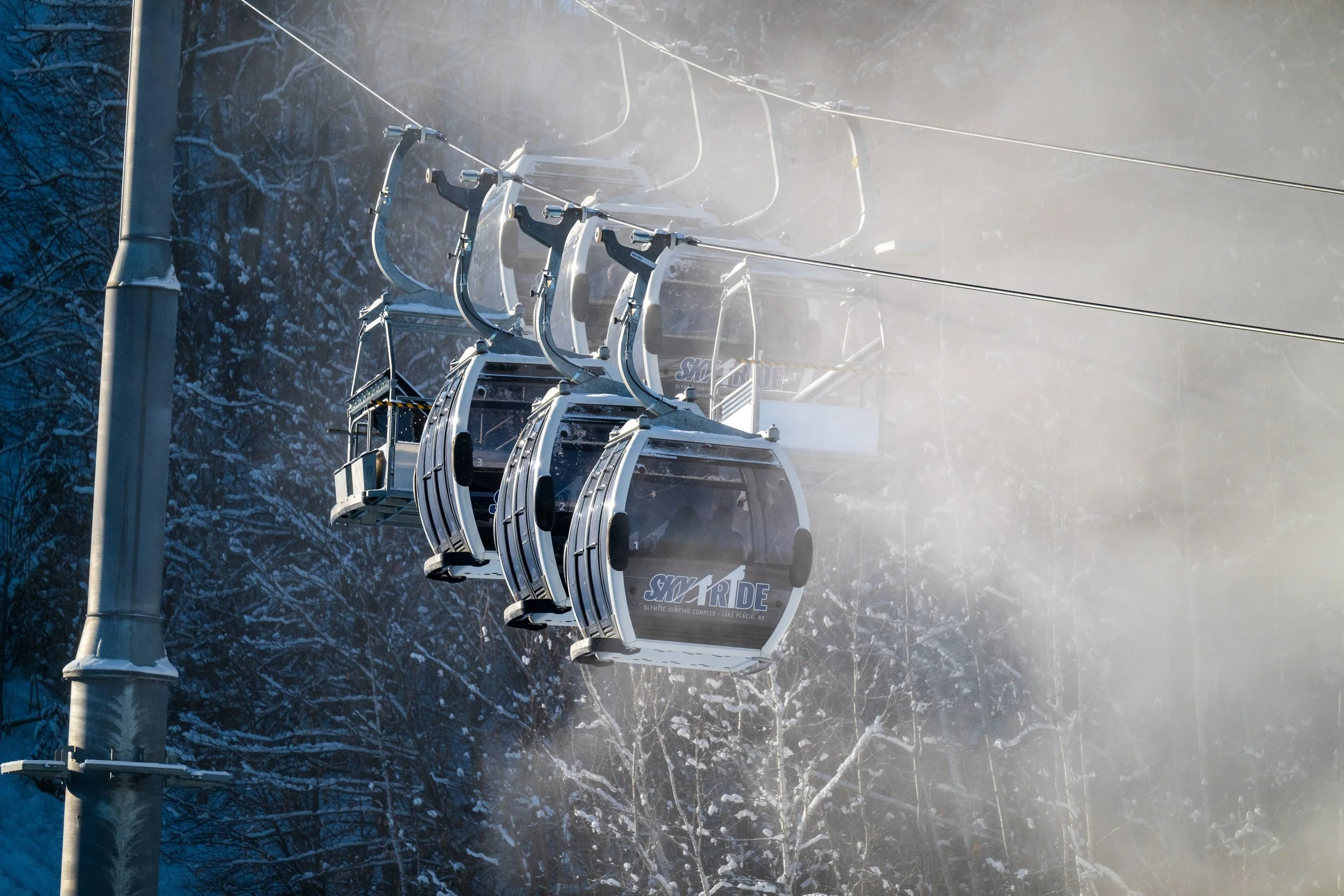 Multiple ski lift chairs suspended from a cable in a snowy, wooded area.
