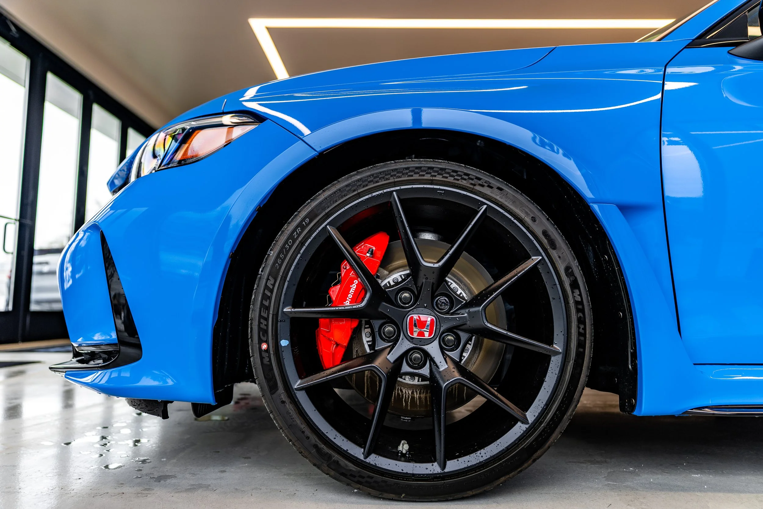 Close-up of the front wheel of a blue Honda car with a black alloy rim, red brake caliper, and Michelin tire, inside a modern showroom.