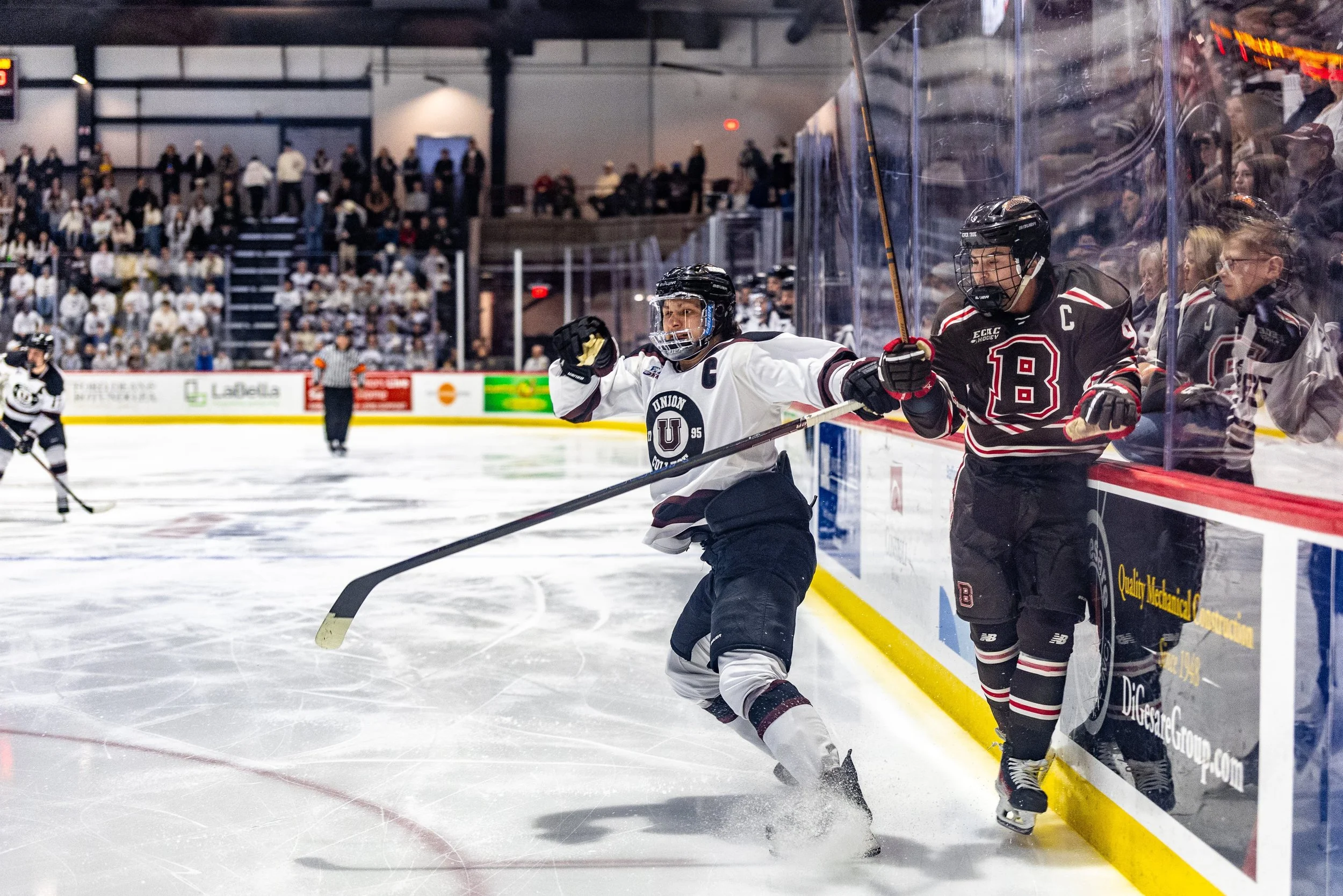 Ice hockey game with two players fighting for puck near the boards, one in white jersey and the other in black jersey, with spectators in the background.