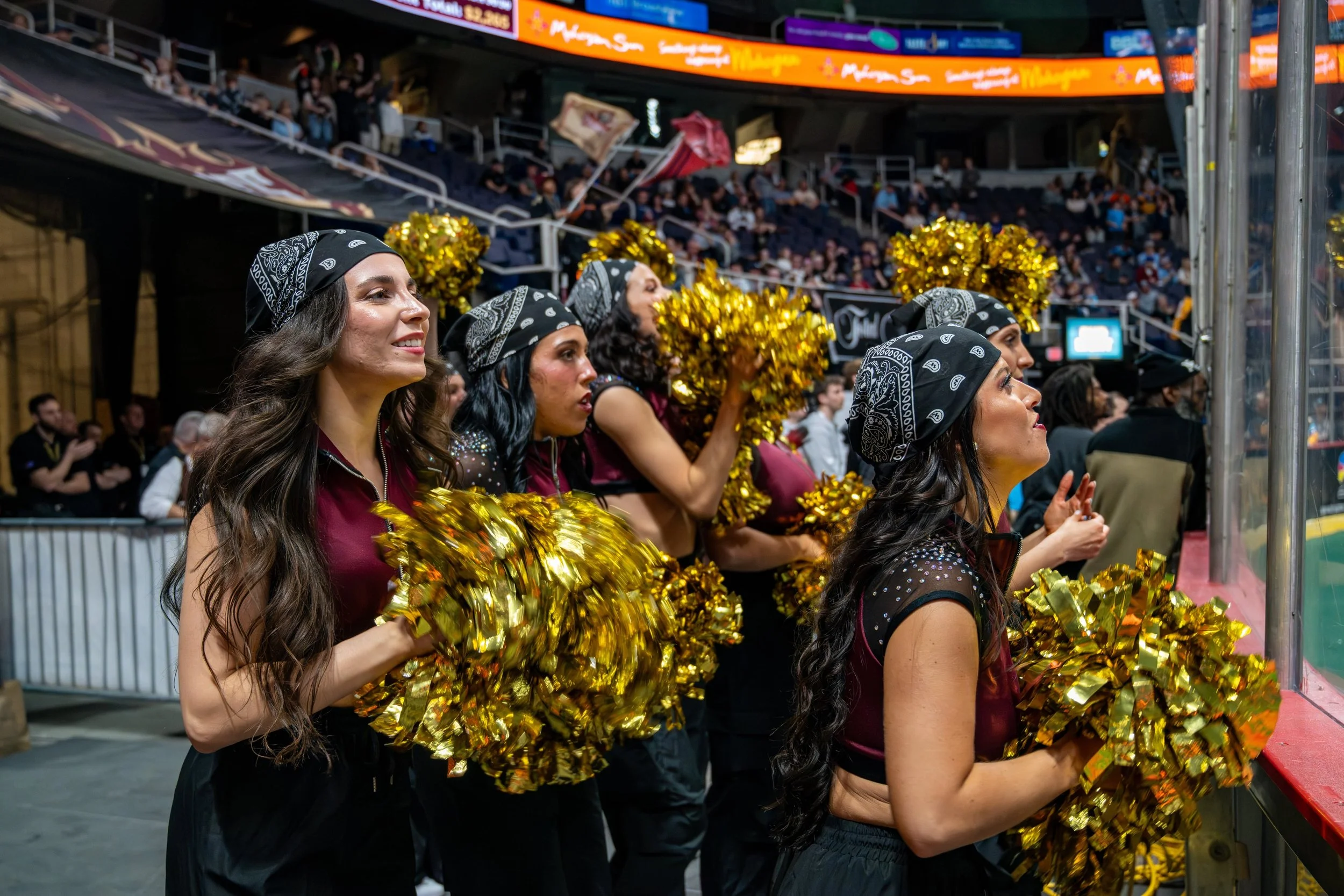 Cheerleaders wearing black bandanas and maroon uniforms holding gold pom-poms and watching a hockey game in an arena.