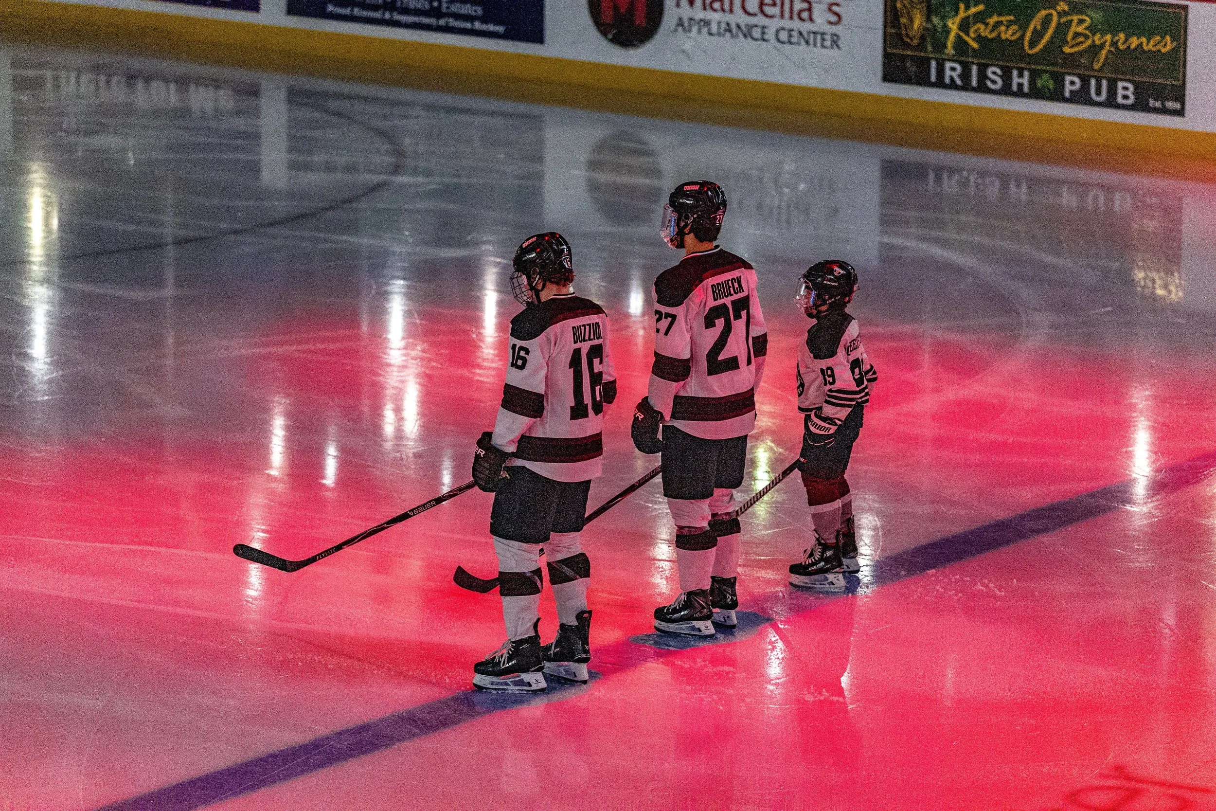 Three young hockey players in black and white jerseys on the ice rink during a game or practice, with advertisements visible in the background.