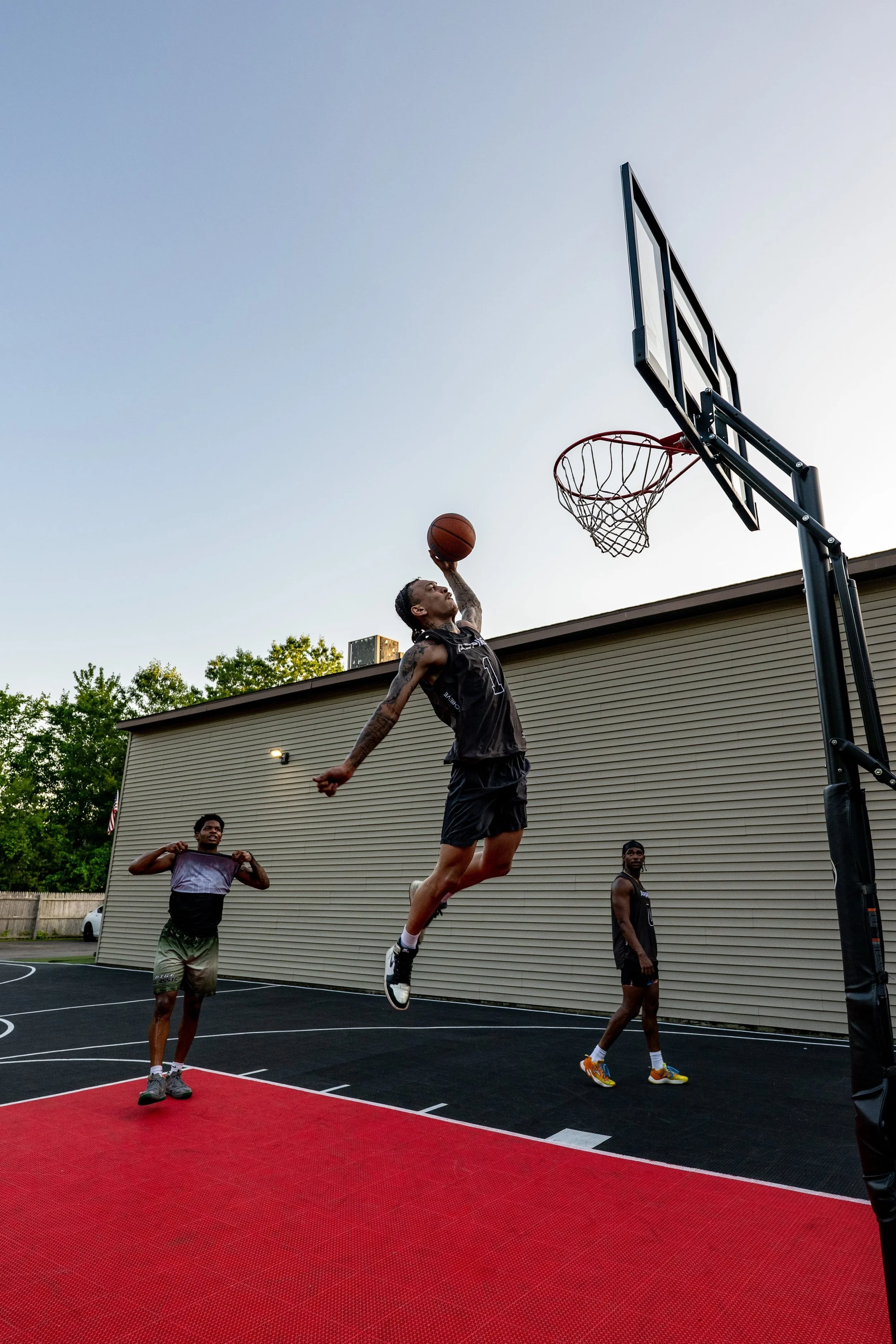 A basketball player jumping towards the hoop in an outdoor court, with two others watching.